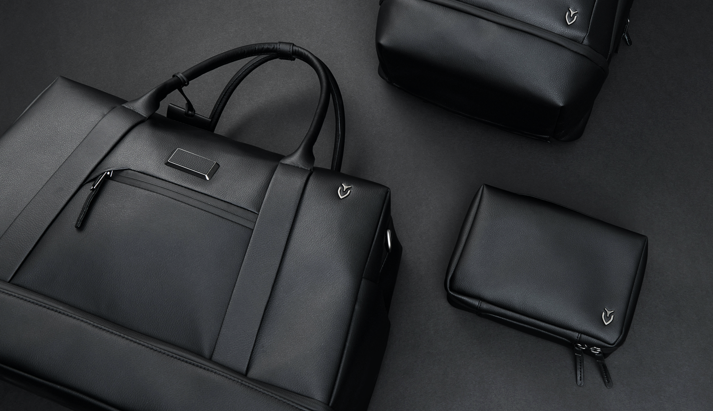 Three black leather handbags of different sizes and a black wallet on a dark surface. The handbags have handles and zipper closures, with small metallic logos on the upper corner.