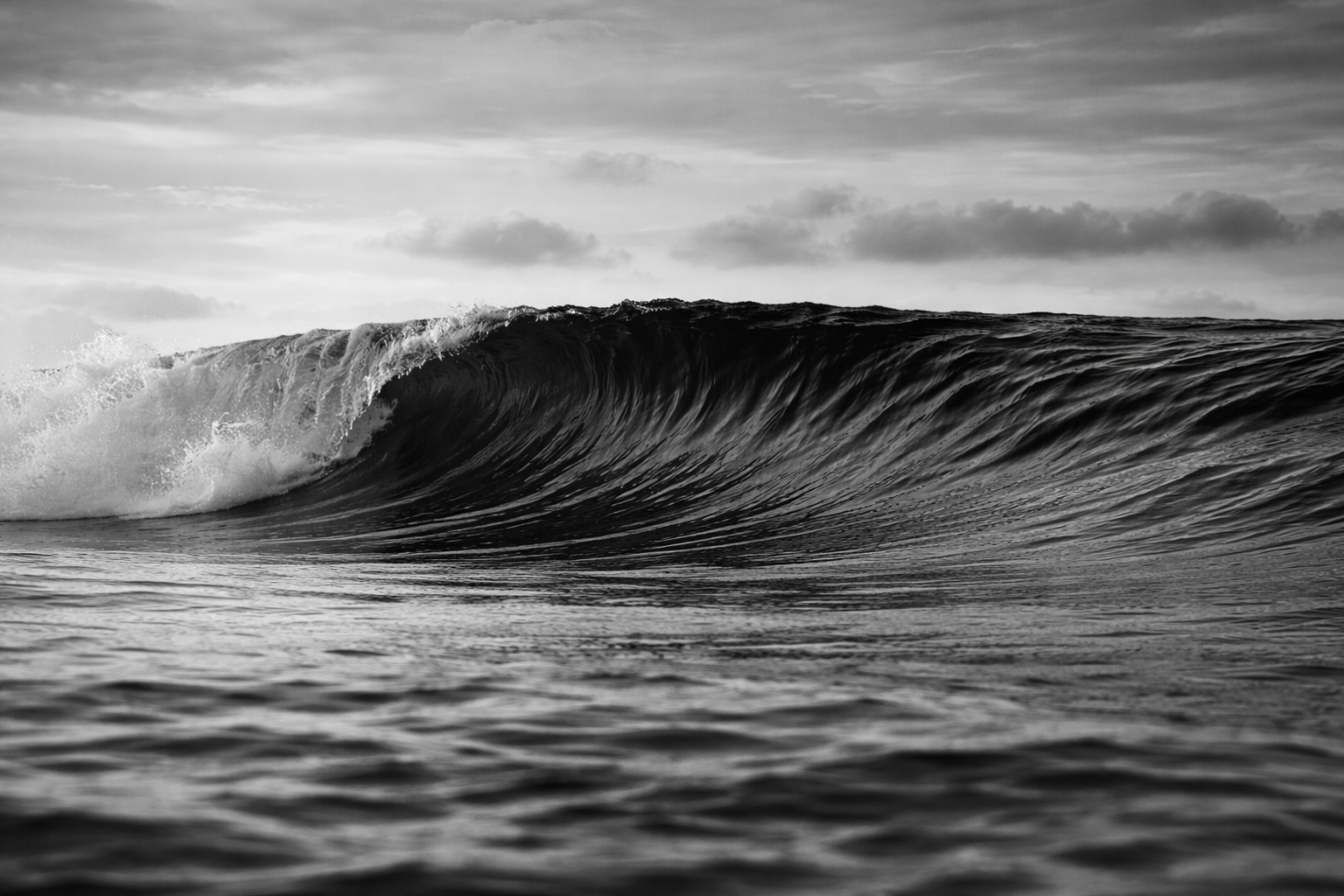 Black and white photograph of an ocean wave curling near the shore, with a cloudy sky in the background.