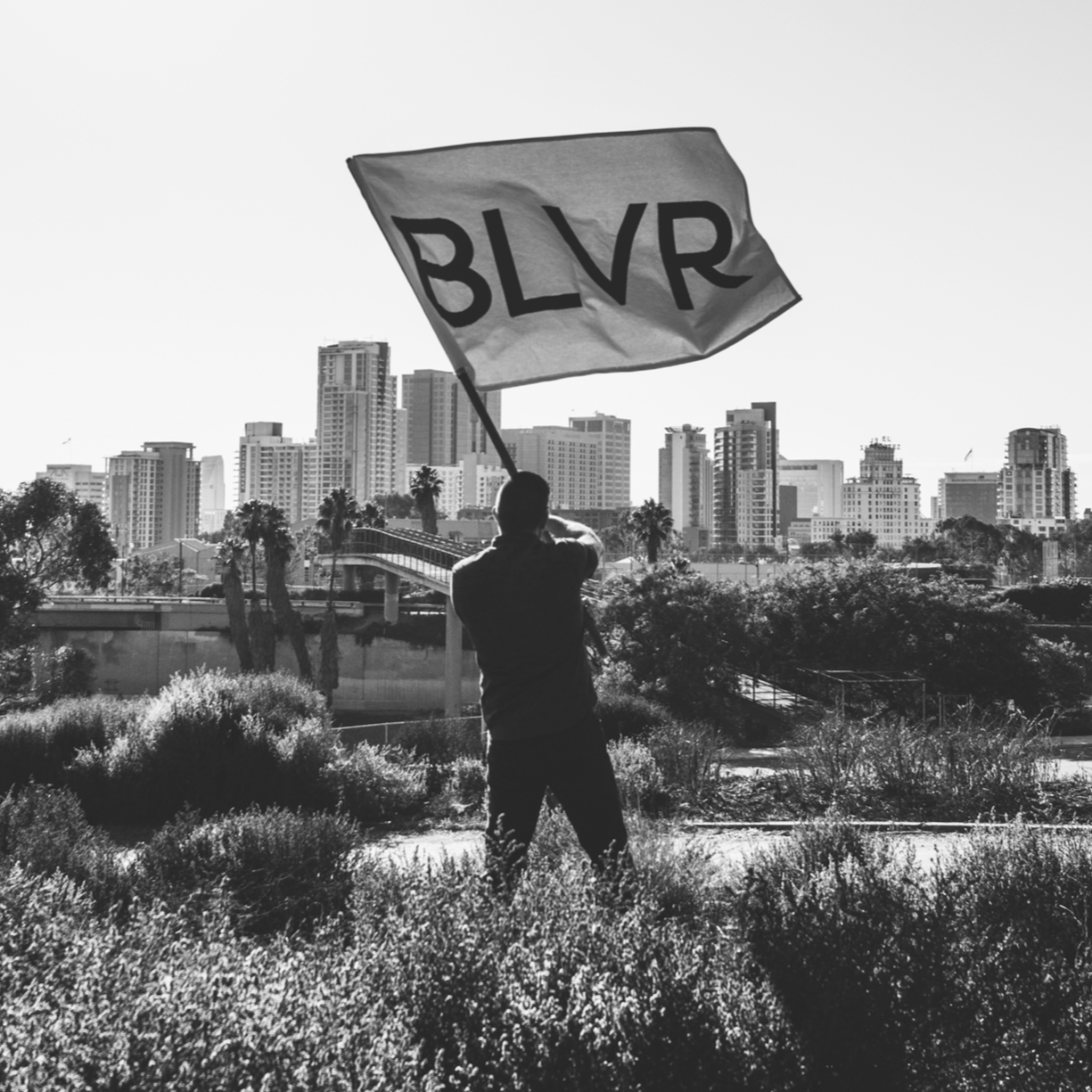 A person holding a flag with the letters 'BLVR' in front of a cityscape with tall buildings and palm trees, in black and white.
