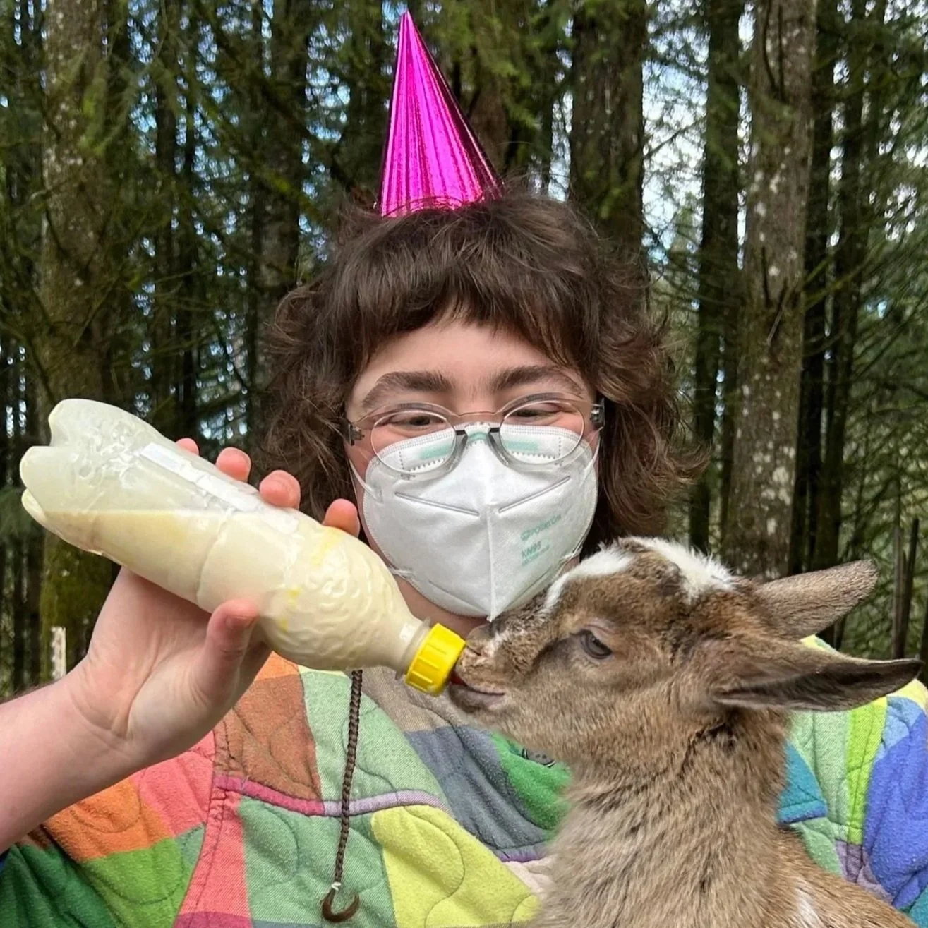 Simka, a person with short curly brown hair and glasses looks at the camera. They are outside with green foliage in the background.