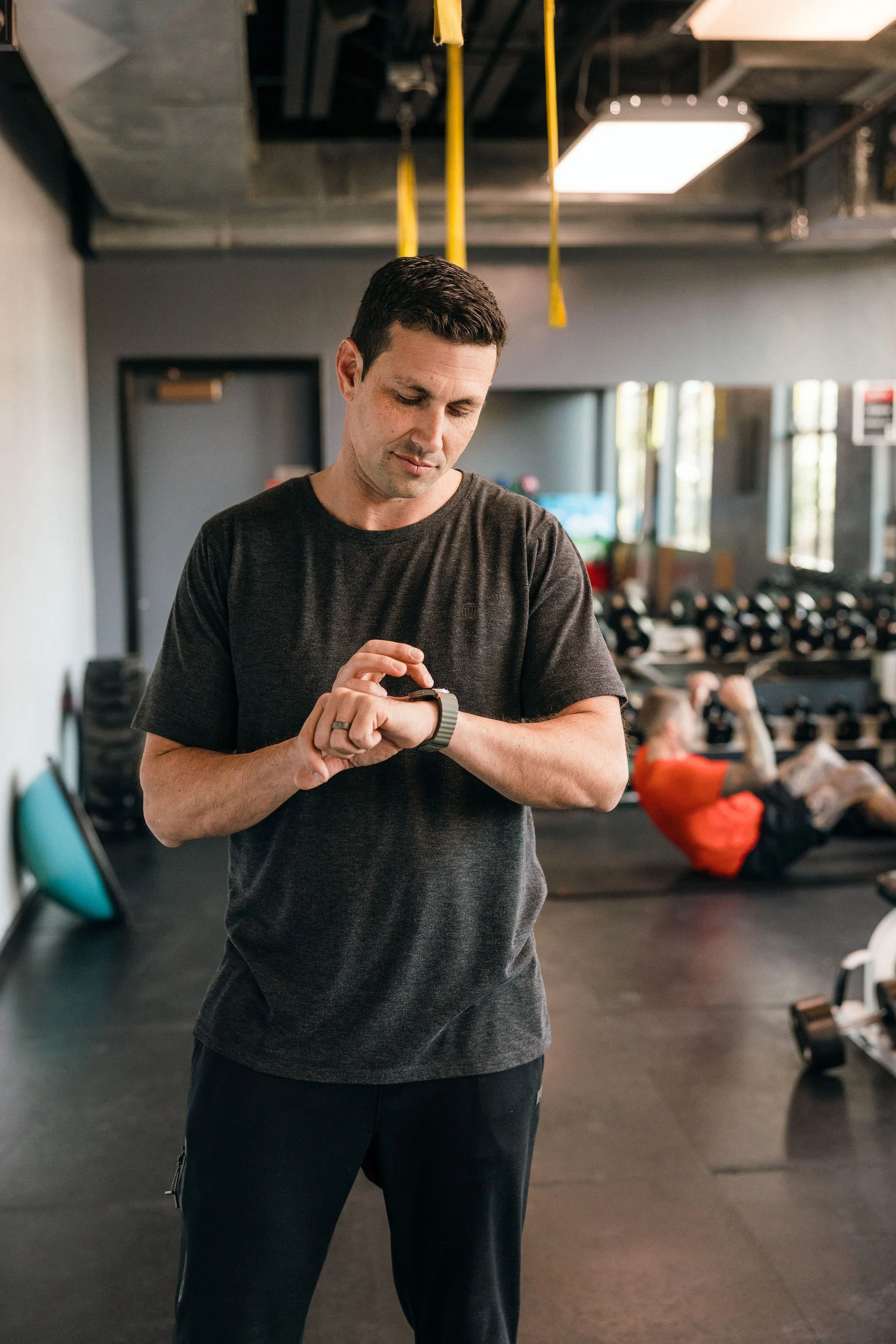 Man in a gym checking a smartwatch as part of a disciplined physical practice connected to 100xLife.
