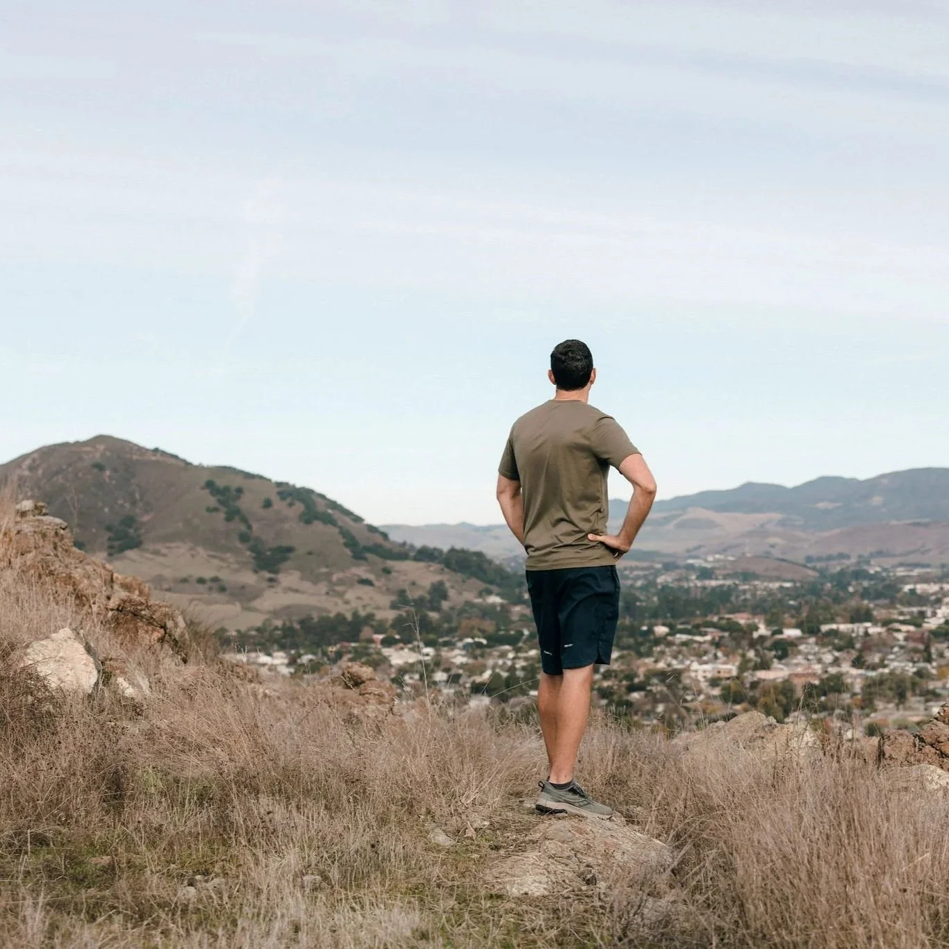 A man standing on a hilltop overlooking a valley and mountains, with his hands on his hips, wearing a brown t-shirt, black shorts, and hiking shoes.
