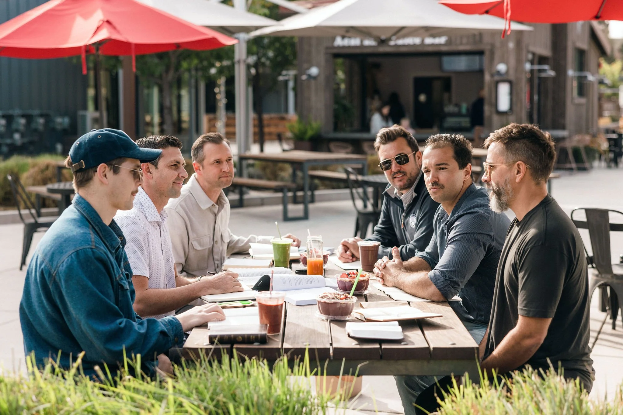 Group of six men sitting at a wooden outdoor table at a cafe, having a discussion with drinks and snacks, under red umbrellas on a sunny day.