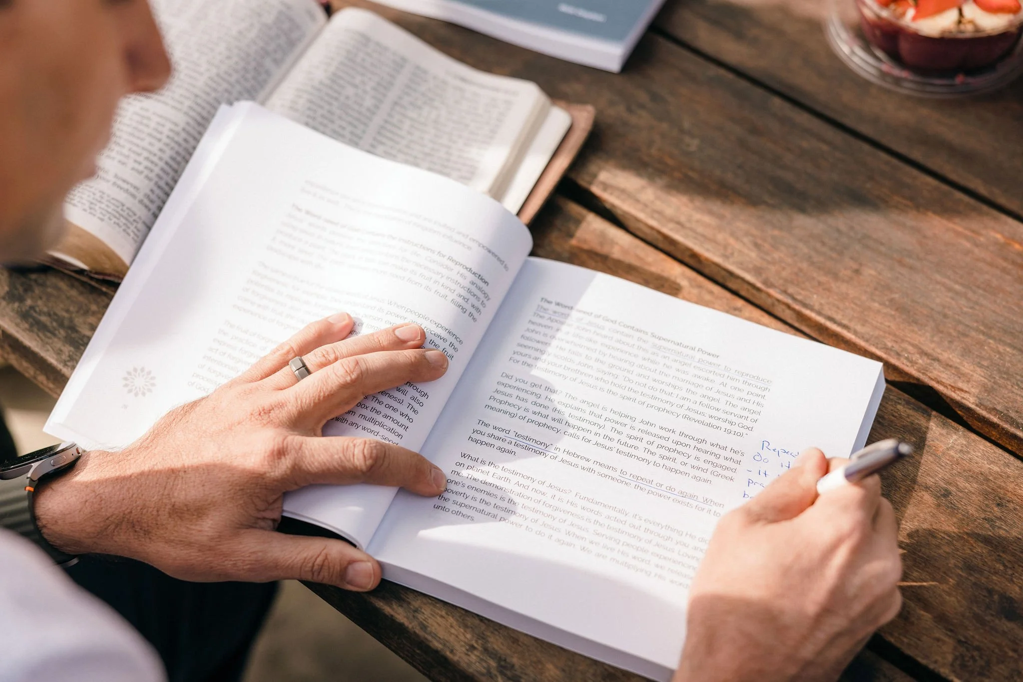 A person pointing at notes in a book with a pen, on a wooden table with other open books and a bowl of mixed fruit.