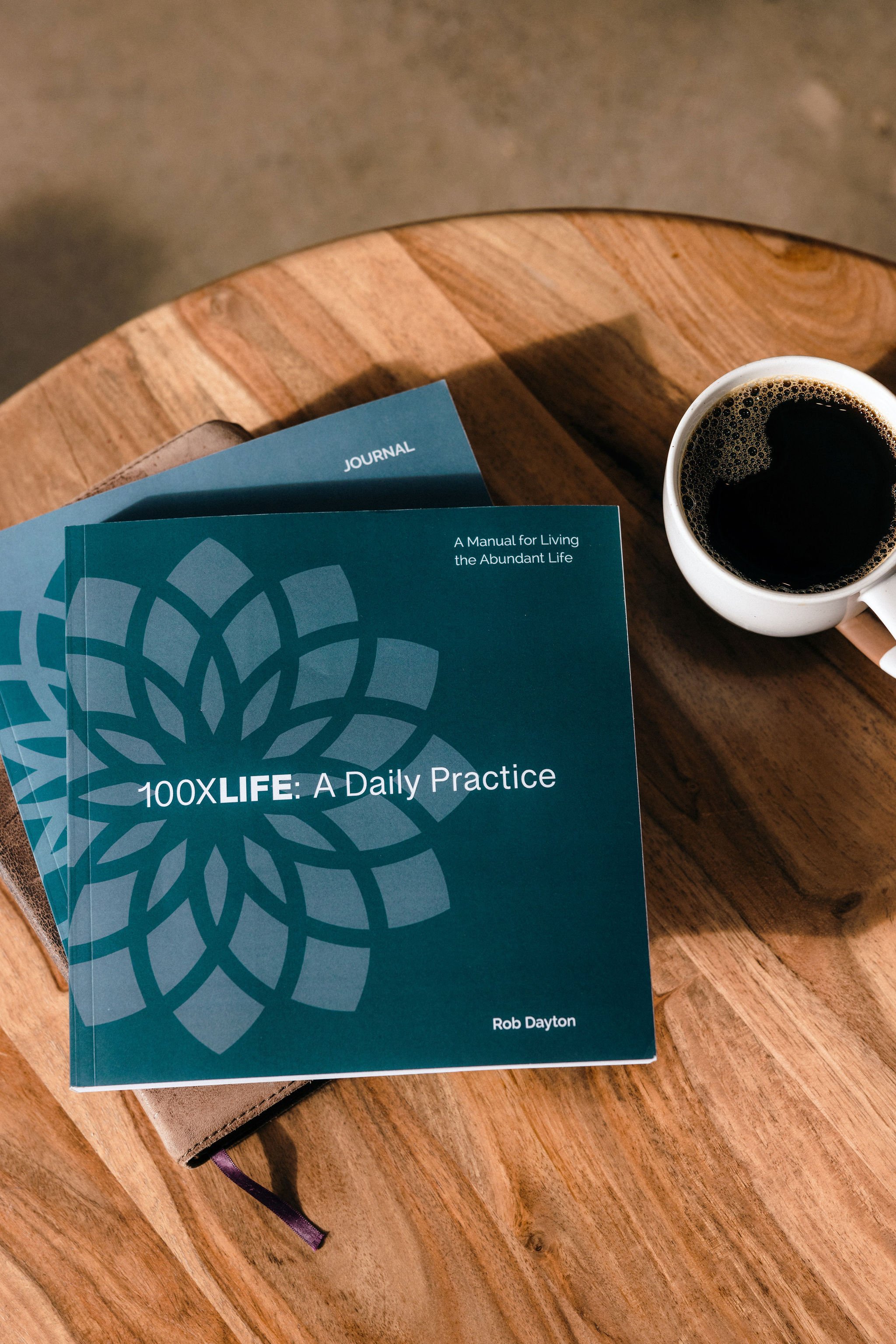 A book titled '100XLife: A Daily Practice' by Rob Dayton and a journal on a wooden table, with a cup of black coffee nearby.