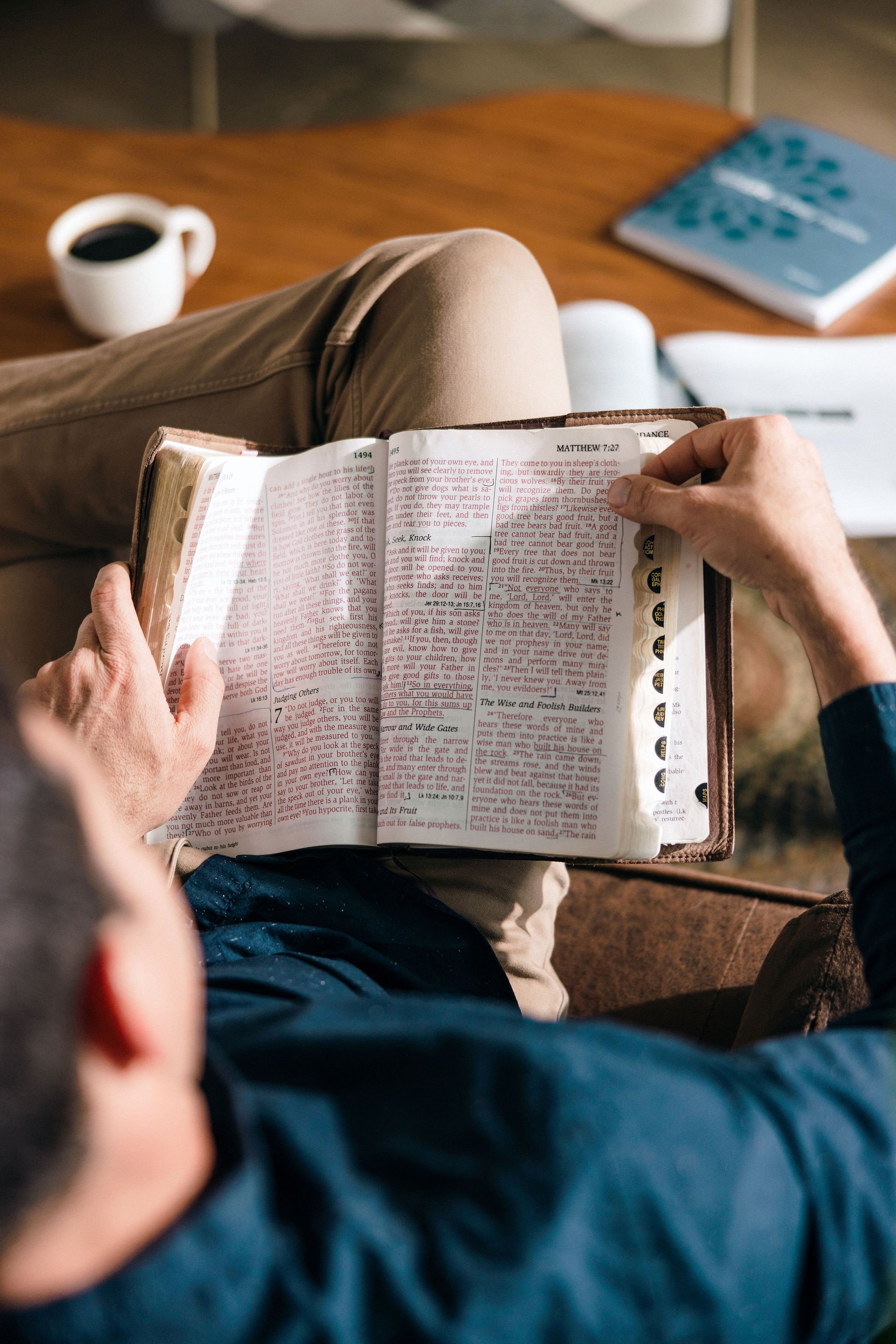 Person reading a Bible with highlighted and underlined passages, sitting on a brown couch with a cup of coffee on a wooden table, a notebook in the background.