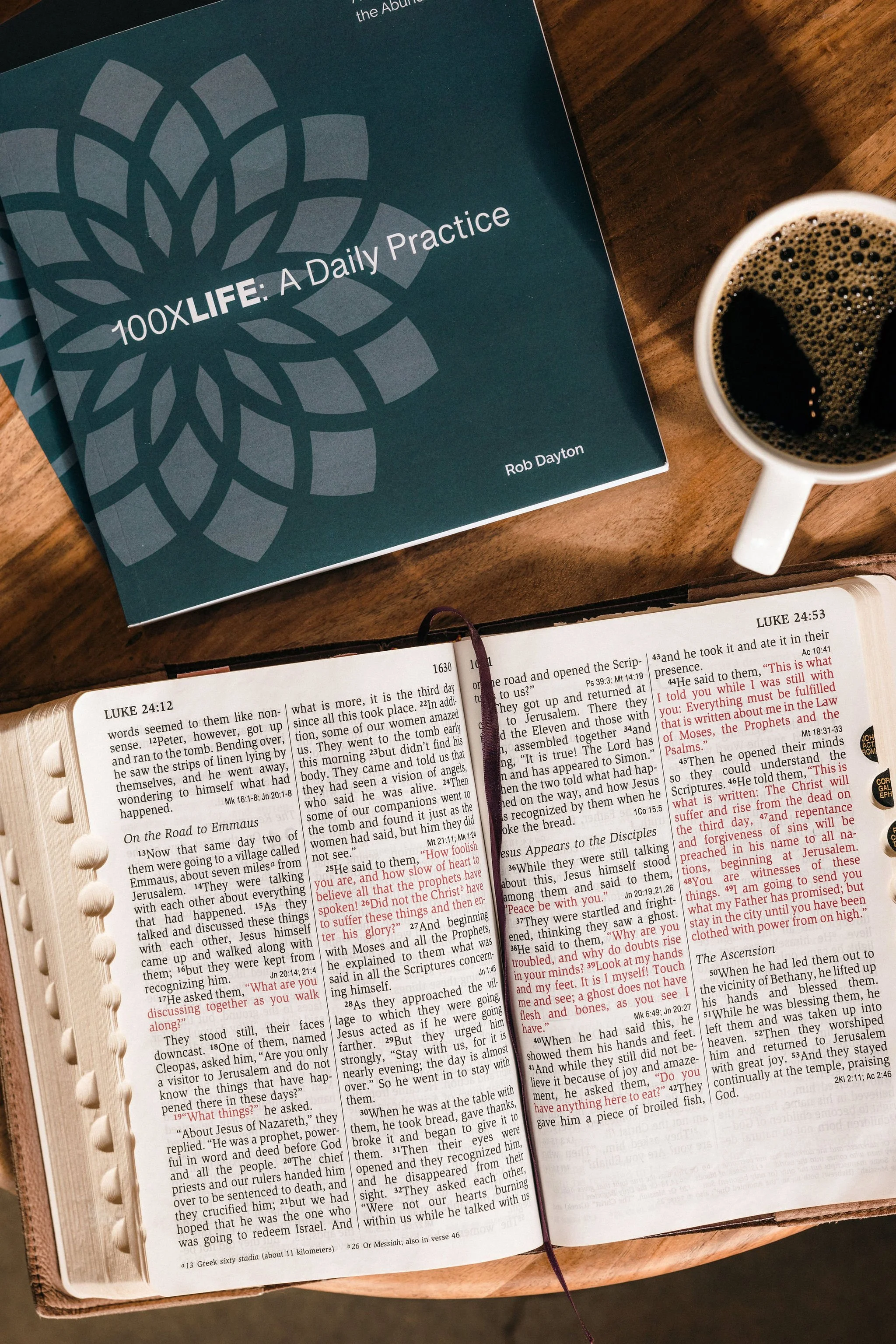 Open Bible with red highlighted text, a cup of coffee, and a book titled '100X LIFE: A Daily Practice' by Rob Dayton on a wooden table.