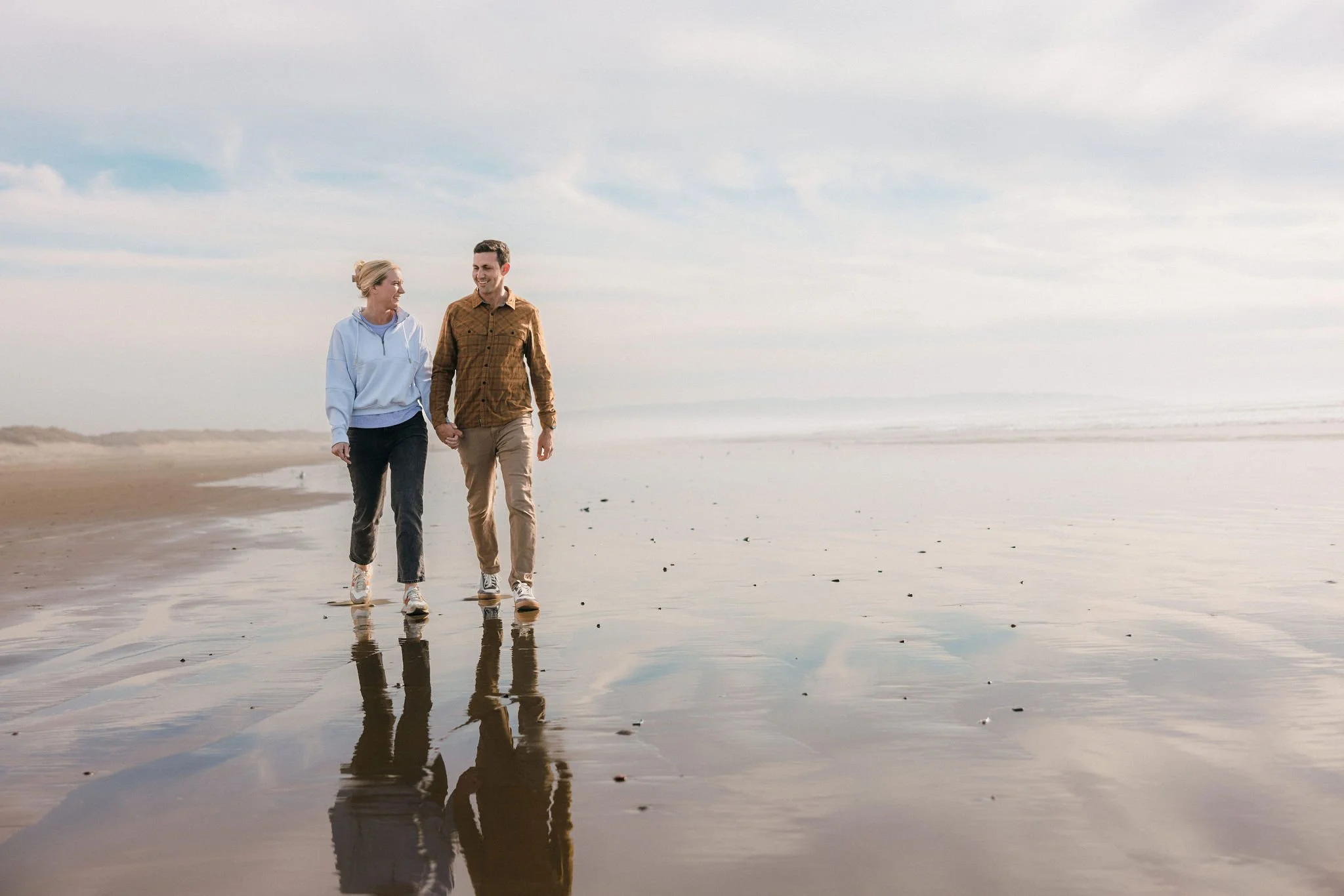 Couple walking together along the beach at sunset, with their reflection visible in the wet sand, representing shared life rhythms and connection.