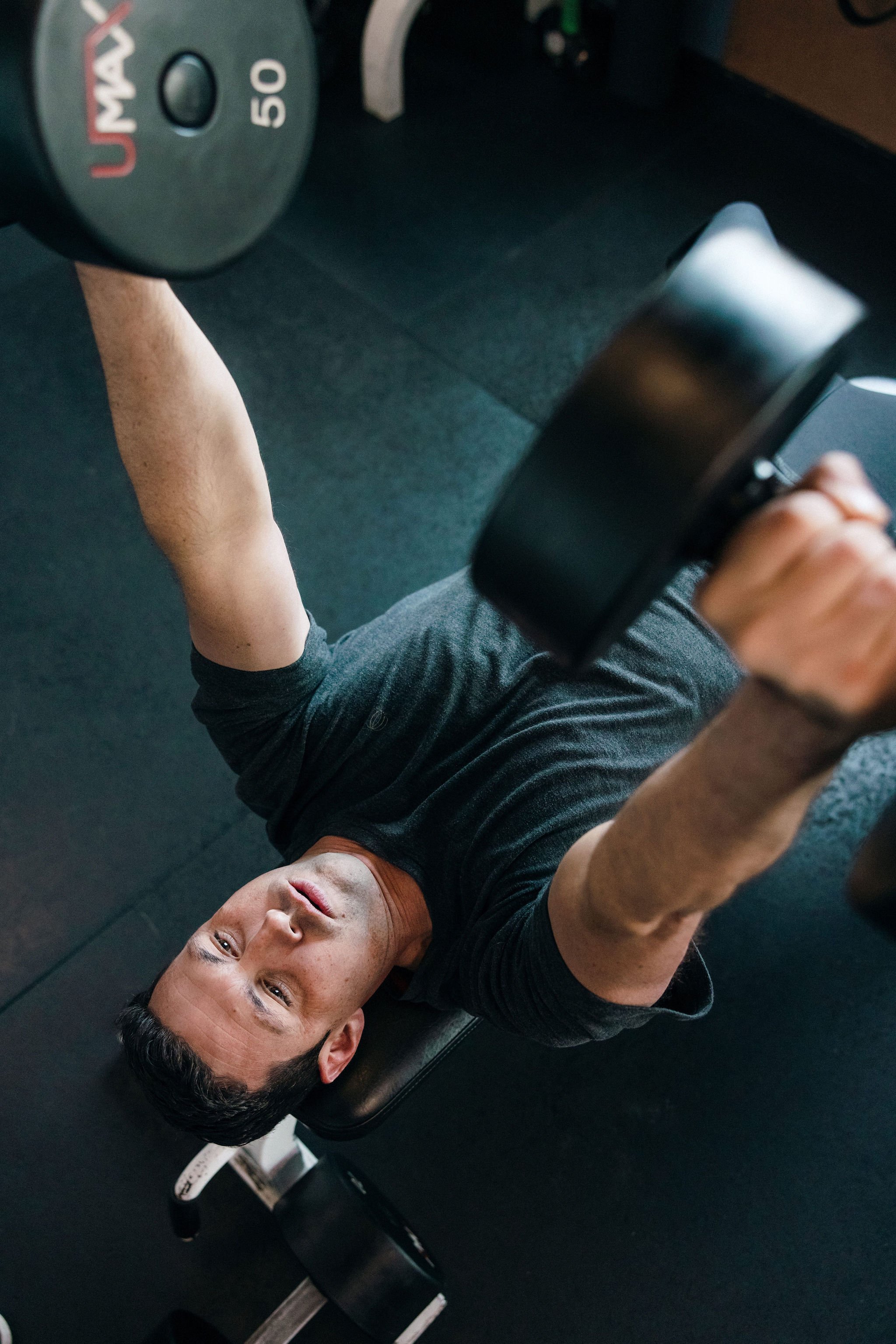 Man lifting a dumbbell in a gym during a disciplined physical practice. He is wearing a black t-shirt and has dark hair. The photo is taken from his perspective, showing the dumbbell and his face.