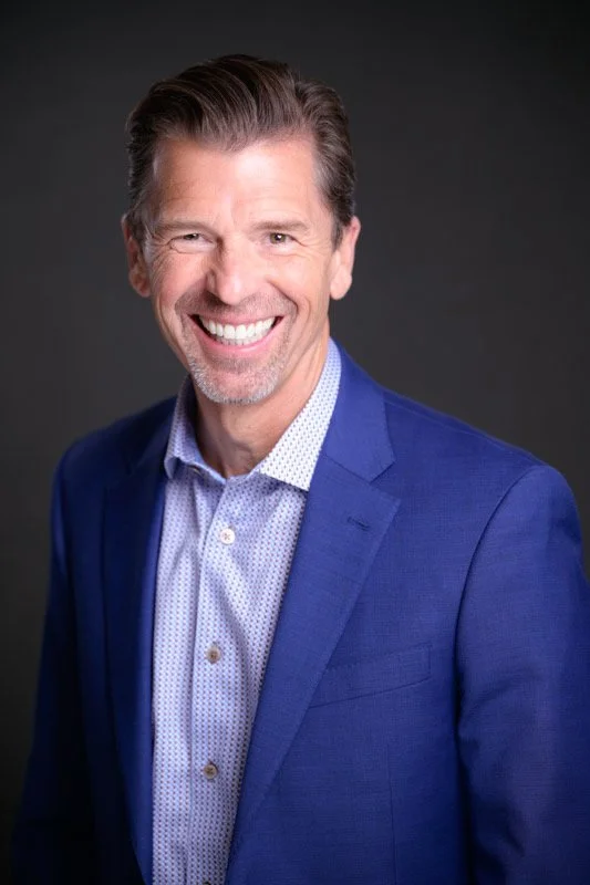 A smiling middle-aged man with short, dark blonde hair and a beard, wearing a blue suit jacket and a white shirt with small blue dots, against a dark background.