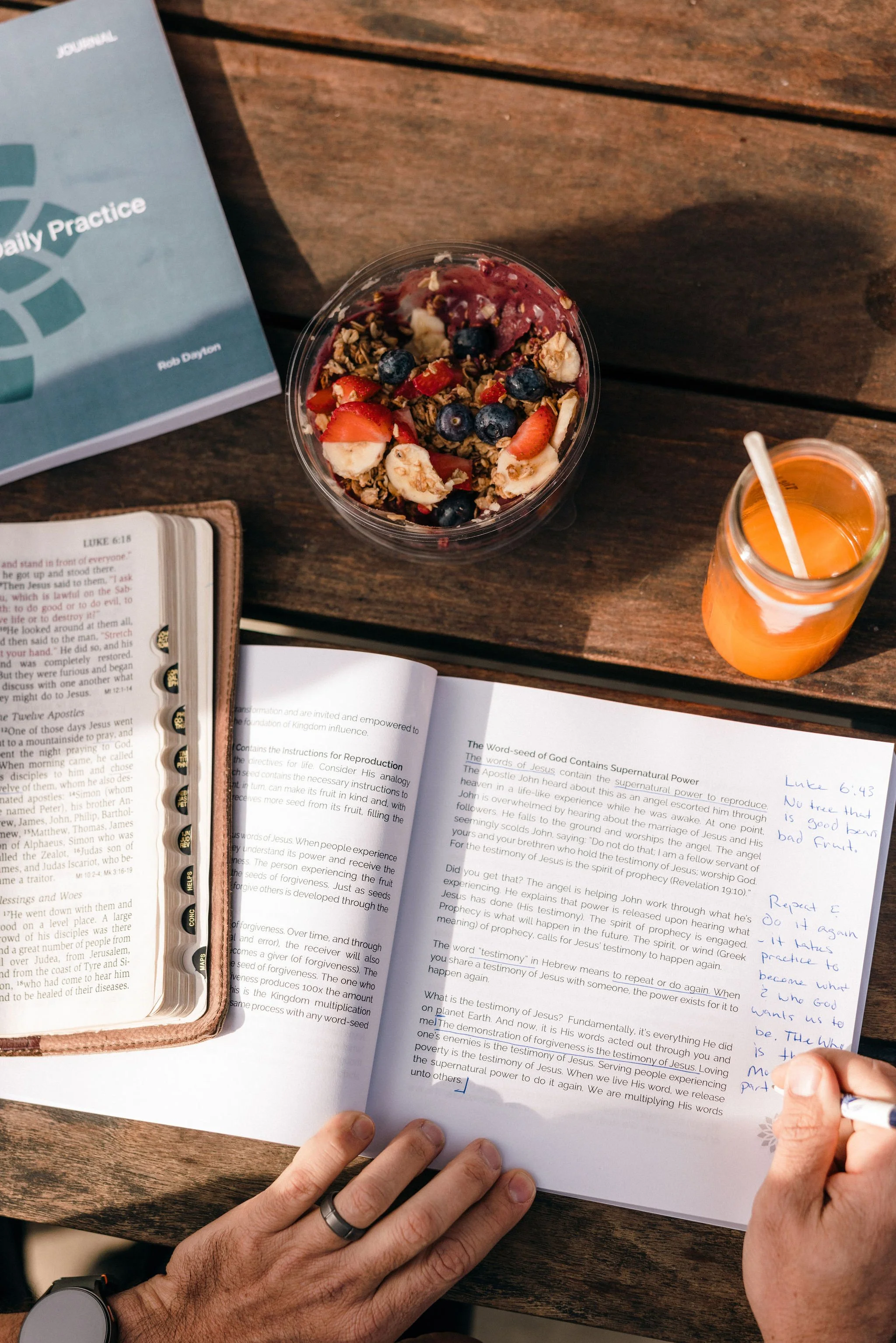 A person studying Bible notes and writing in a notebook during a focused daily practice, with a bowl of yogurt topped with strawberries, blueberries, granola, and bananas, and a jar of orange juice on a wooden table.