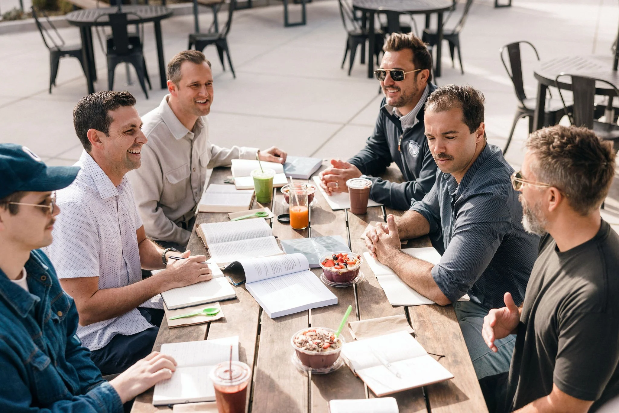 Six men gathered around a table in conversation representing community and shared practice in 100xLife.