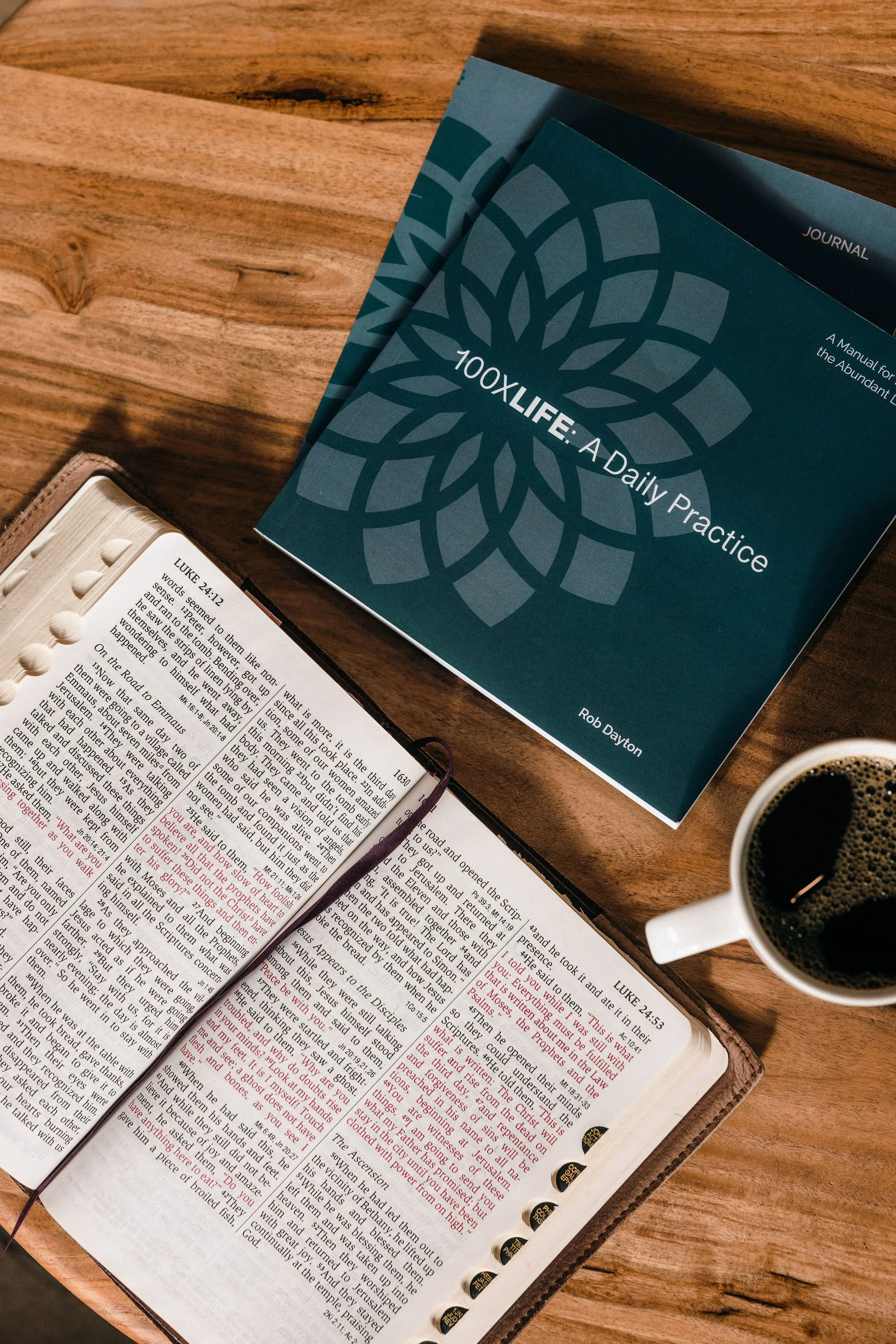 Open Bible, journal, coffee, and the "100xLIFE: A Daily Practice" book arranged for Scripture-based reflection.