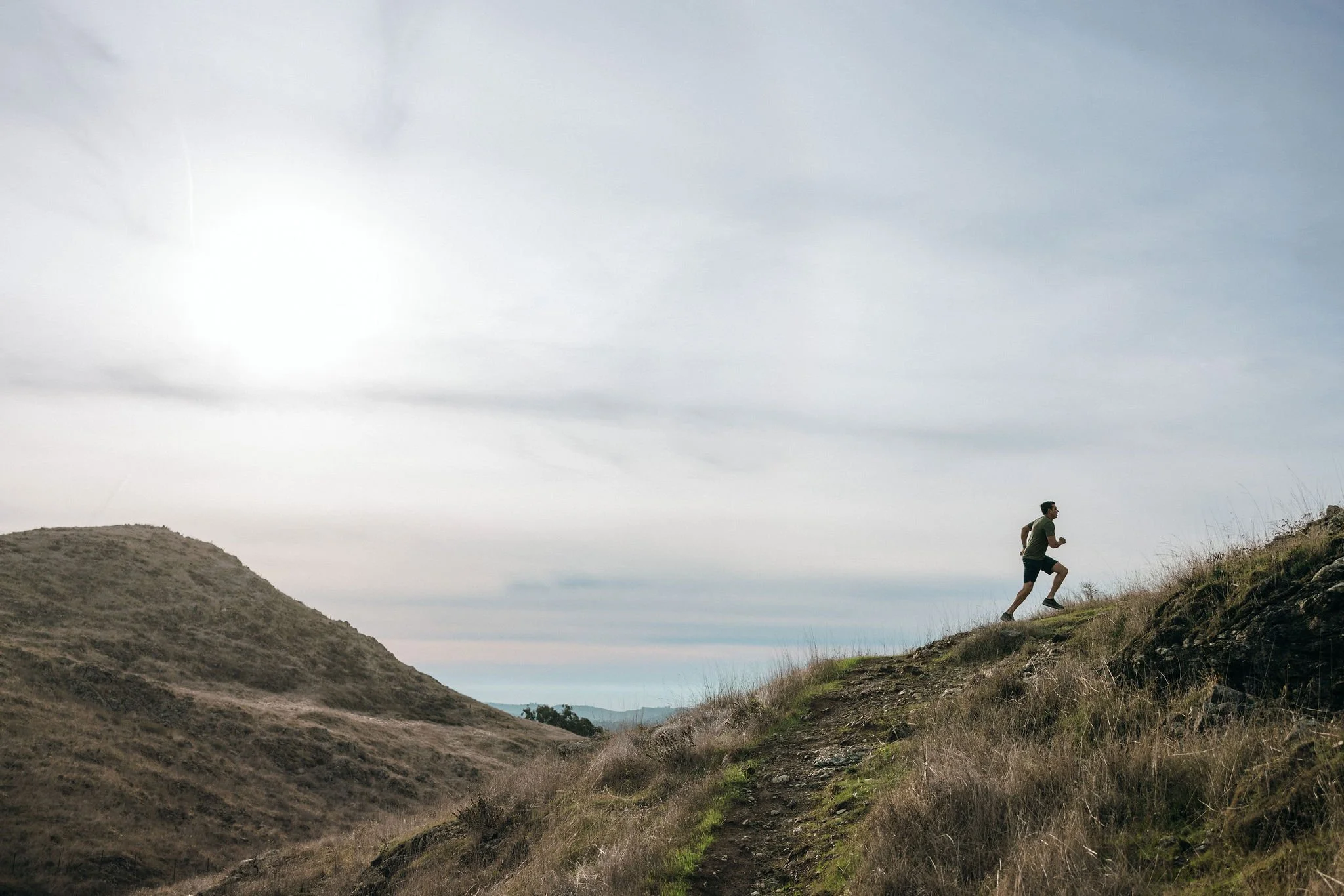 A man running up a dirt trail on a hillside with dry grass and shrubs, under a cloudy sky with the sun partially hidden.