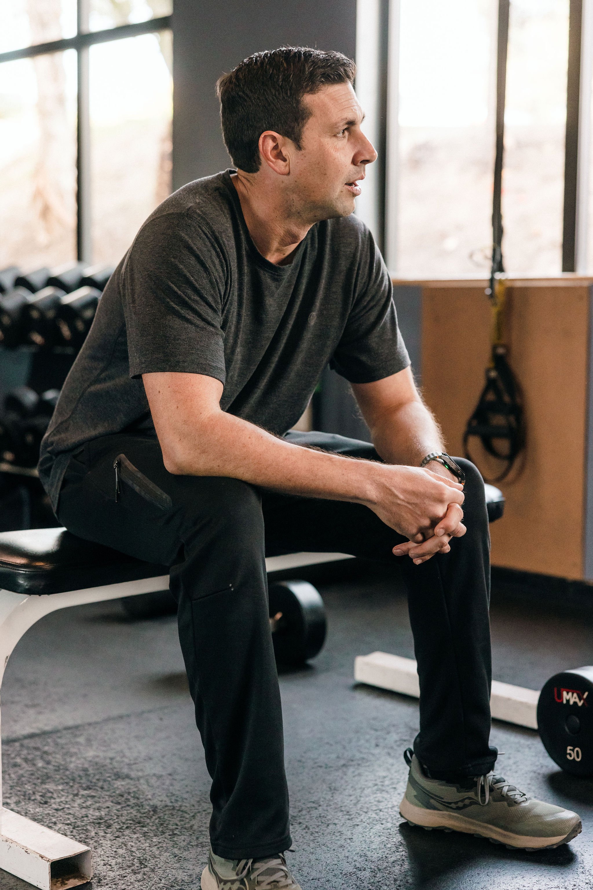 A man with dark hair, wearing a gray t-shirt, black pants, and gray athletic shoes, sits on a workout bench in a gym, reflecting during a daily discipline tied to body, mind, and spirit.