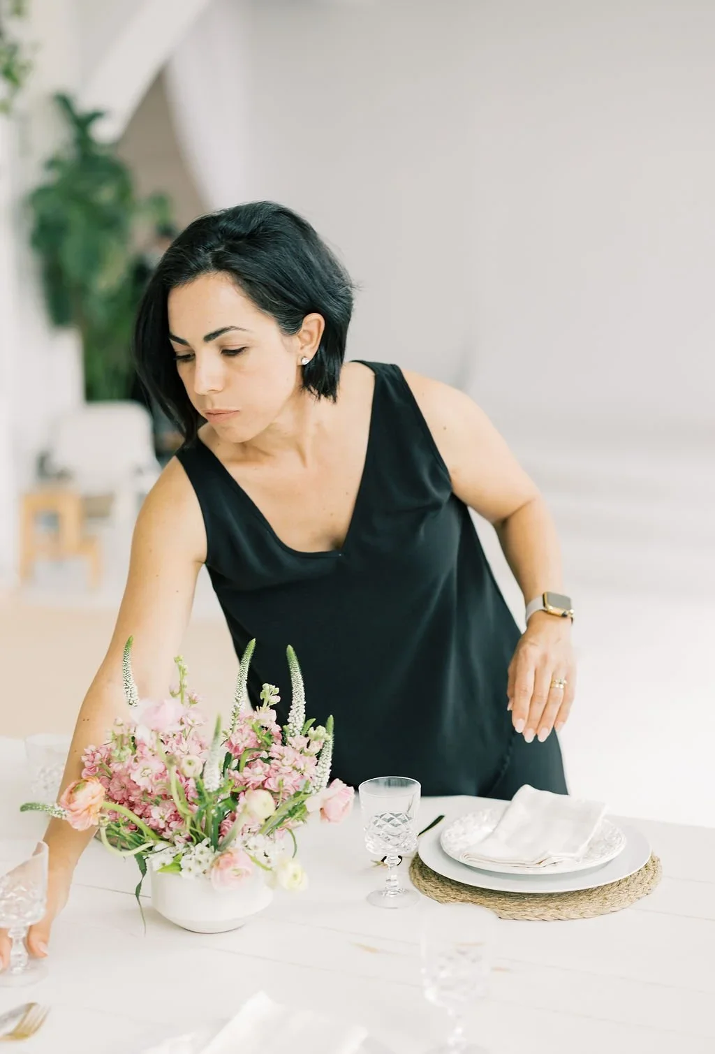 A woman with black hair and a black sleeveless dress arranging a floral centerpiece on a dining table with white plates, glasses, and a woven placemat in a bright room.
