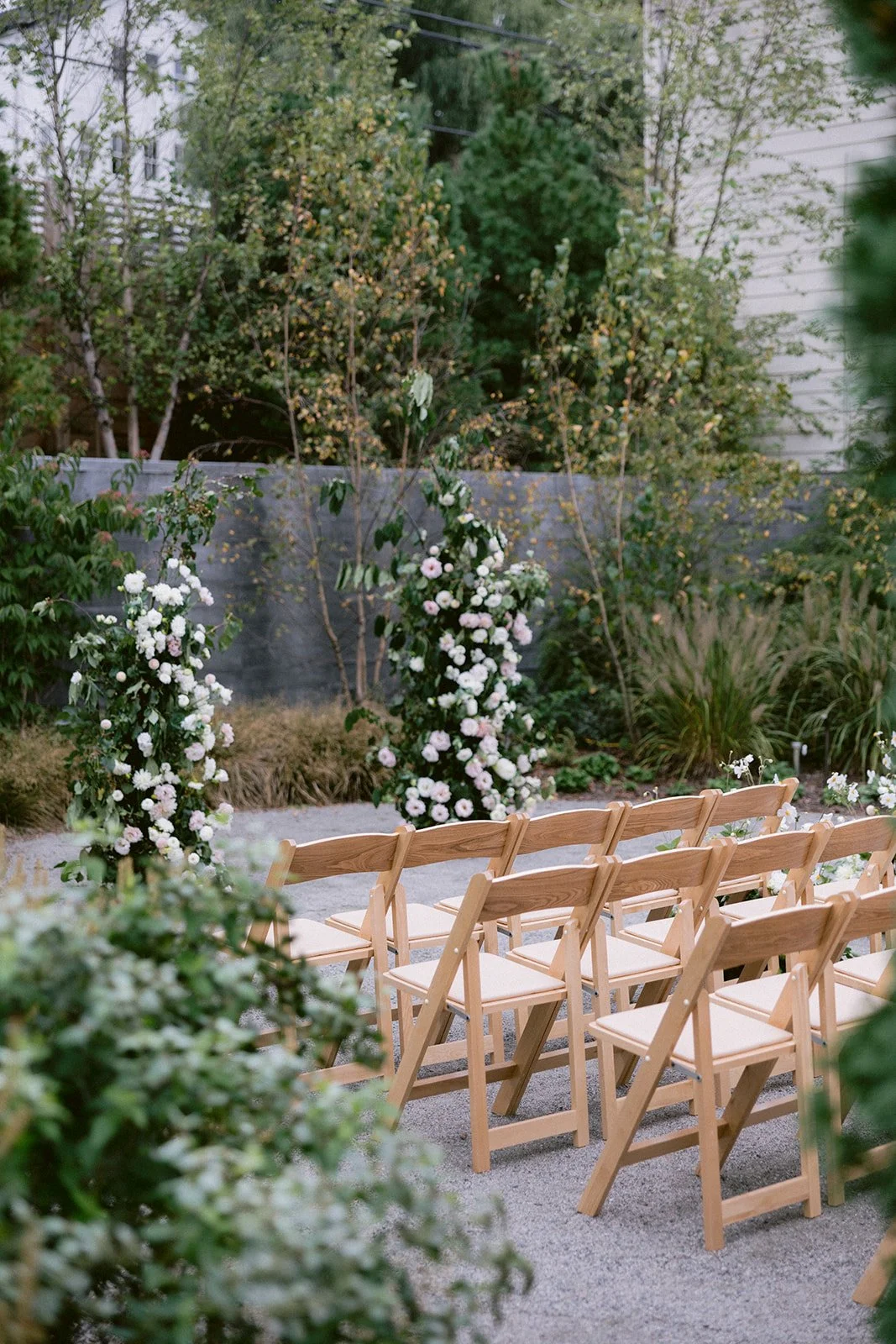 Outdoor wedding ceremony setup with wooden chairs arranged in rows facing floral archways, surrounded by lush greenery and flowers.