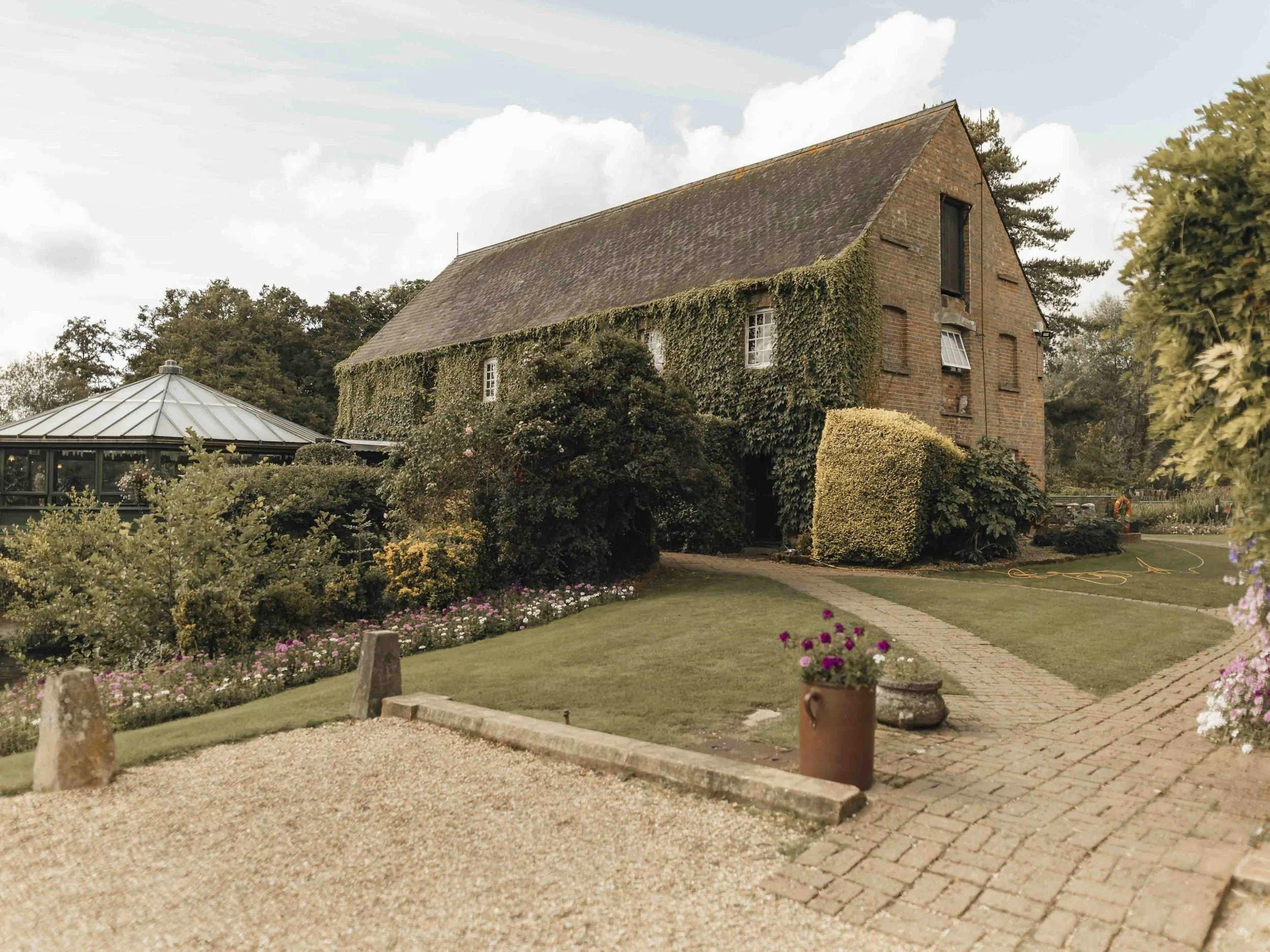 A brick house with ivy growing up its side, surrounded by a well-maintained garden with colorful flowers and a brick pathway.