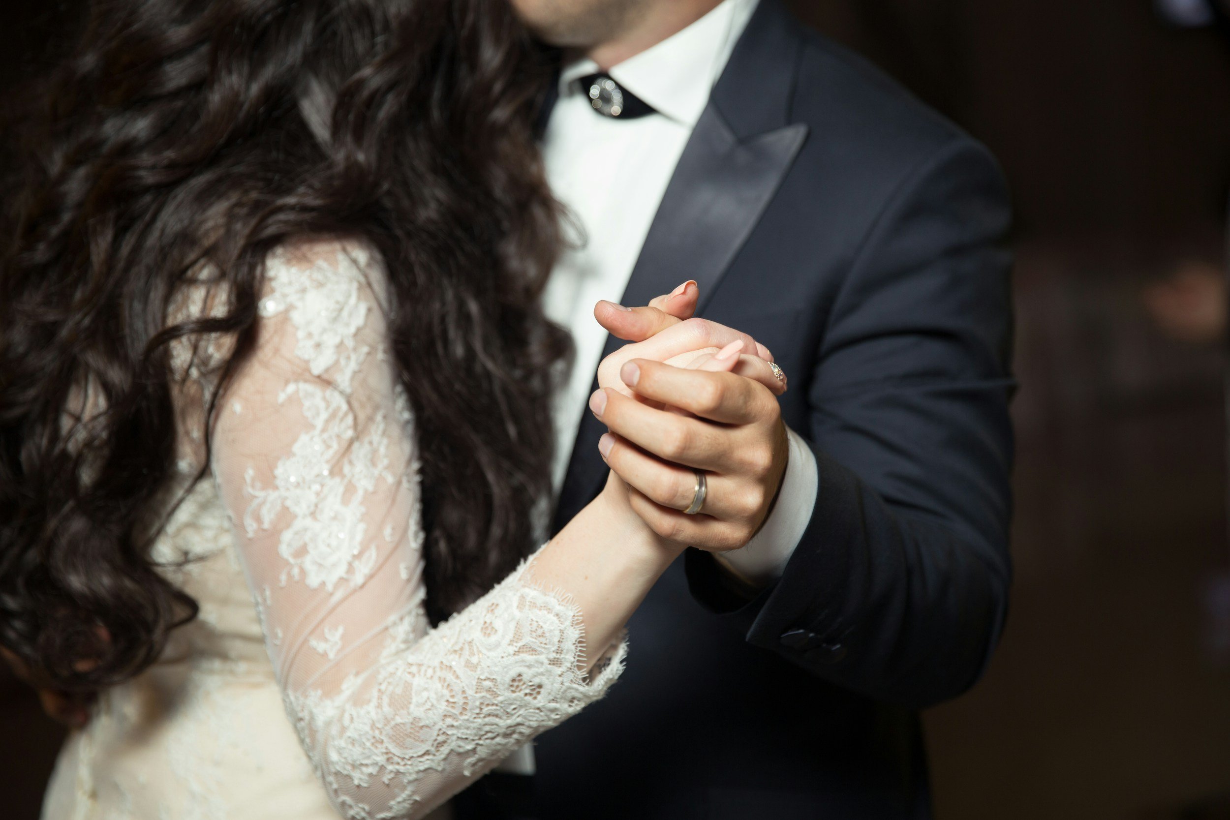 Close-up of a bride and groom holding hands during their wedding dance. The bride's lace sleeve and the groom's formal suit are visible.