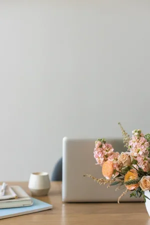 A laptop on a wooden desk with a bouquet of pink and peach flowers in a white vase, a small white candle holder, and some papers and a pen.
