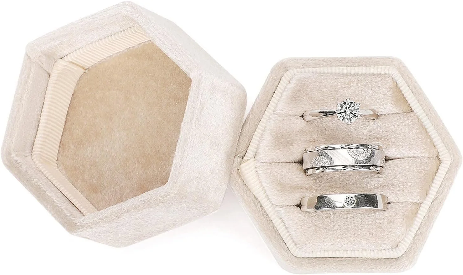 A hexagonal cream-colored jewelry box with three silver rings inside, one with a diamond solitaire, one with an engraved band, and one with a circular design.