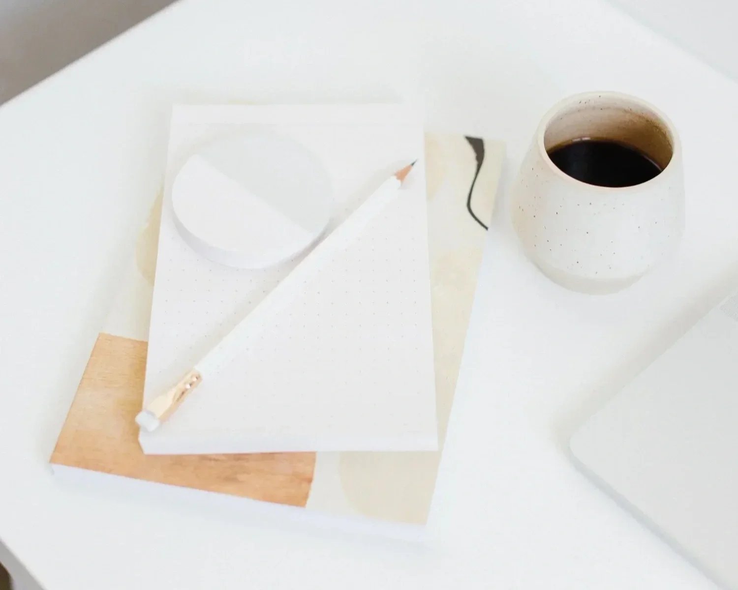 A white table with a white notepad, a pencil, and a white ceramic cup filled with black coffee.