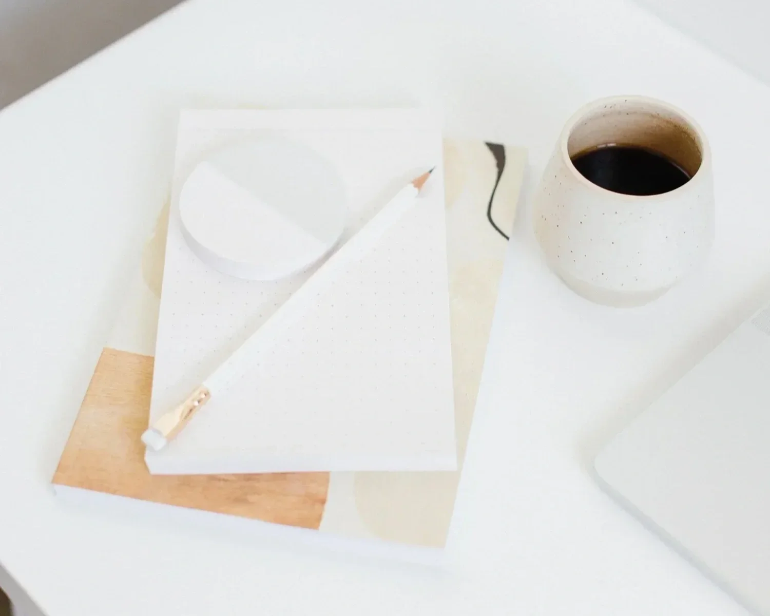 A white table with a white notepad, a pencil, and a white ceramic cup filled with black coffee.