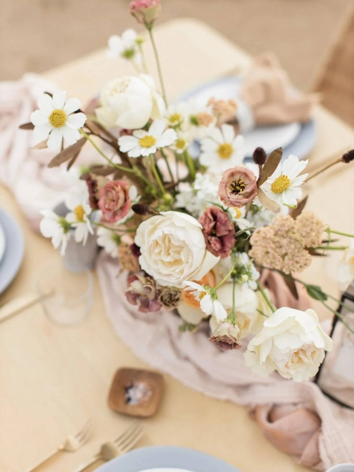 A floral centerpiece with white roses, pink ranunculus, daisies, and other flowers on a table decorated with a soft pink cloth.