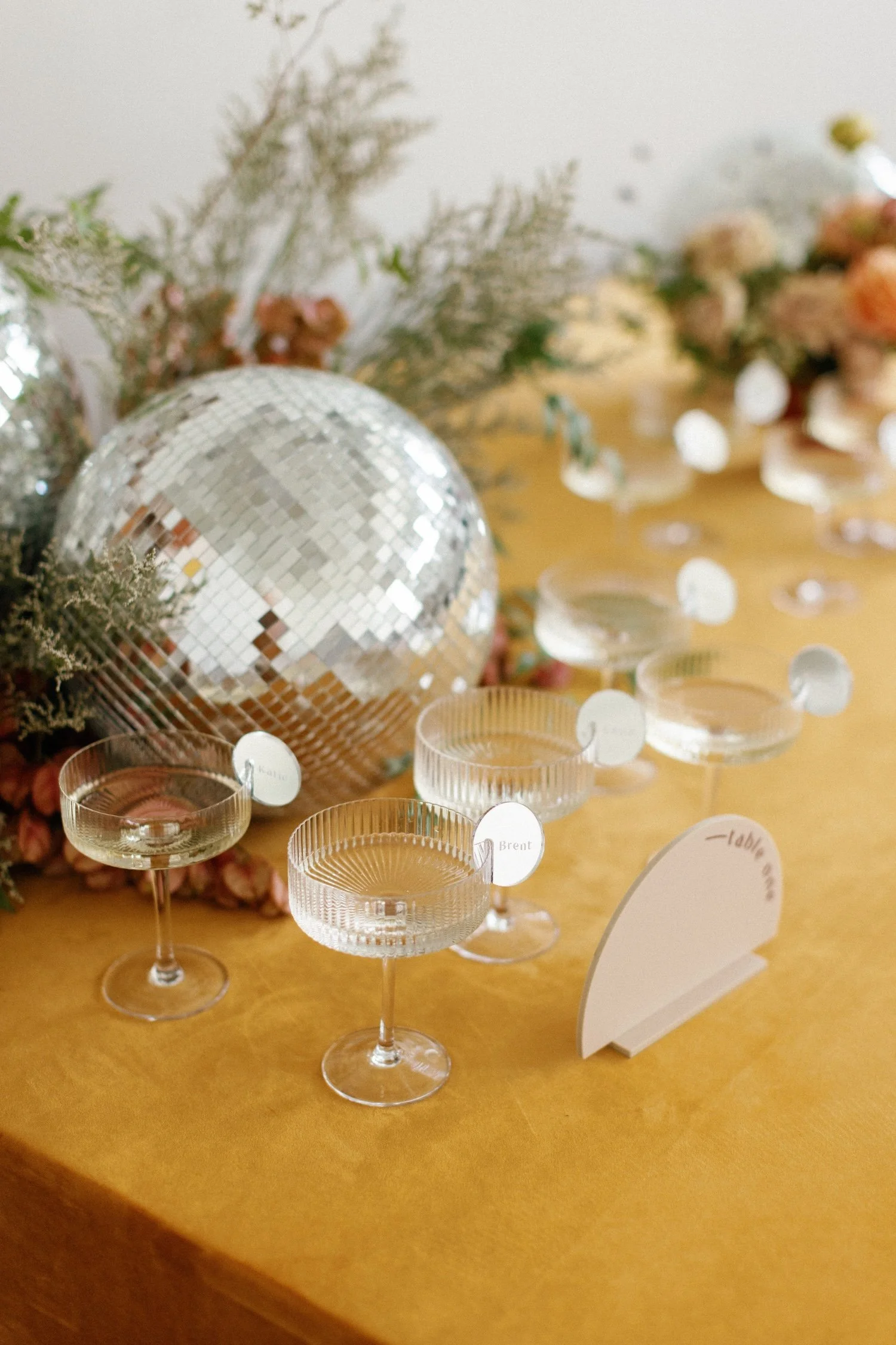 Table decorated with a large disco ball, floral arrangements, and multiple coupe glasses with place cards, on a yellow tablecloth.