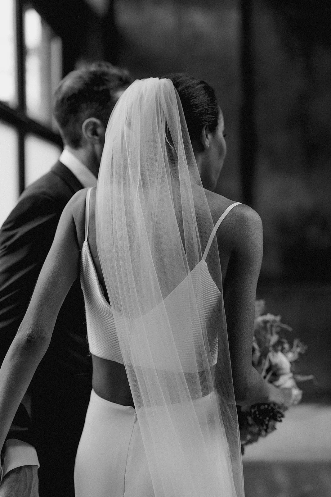 A black-and-white photo of a bride in a wedding dress and veil, standing next to a groom, with other figures in the background.