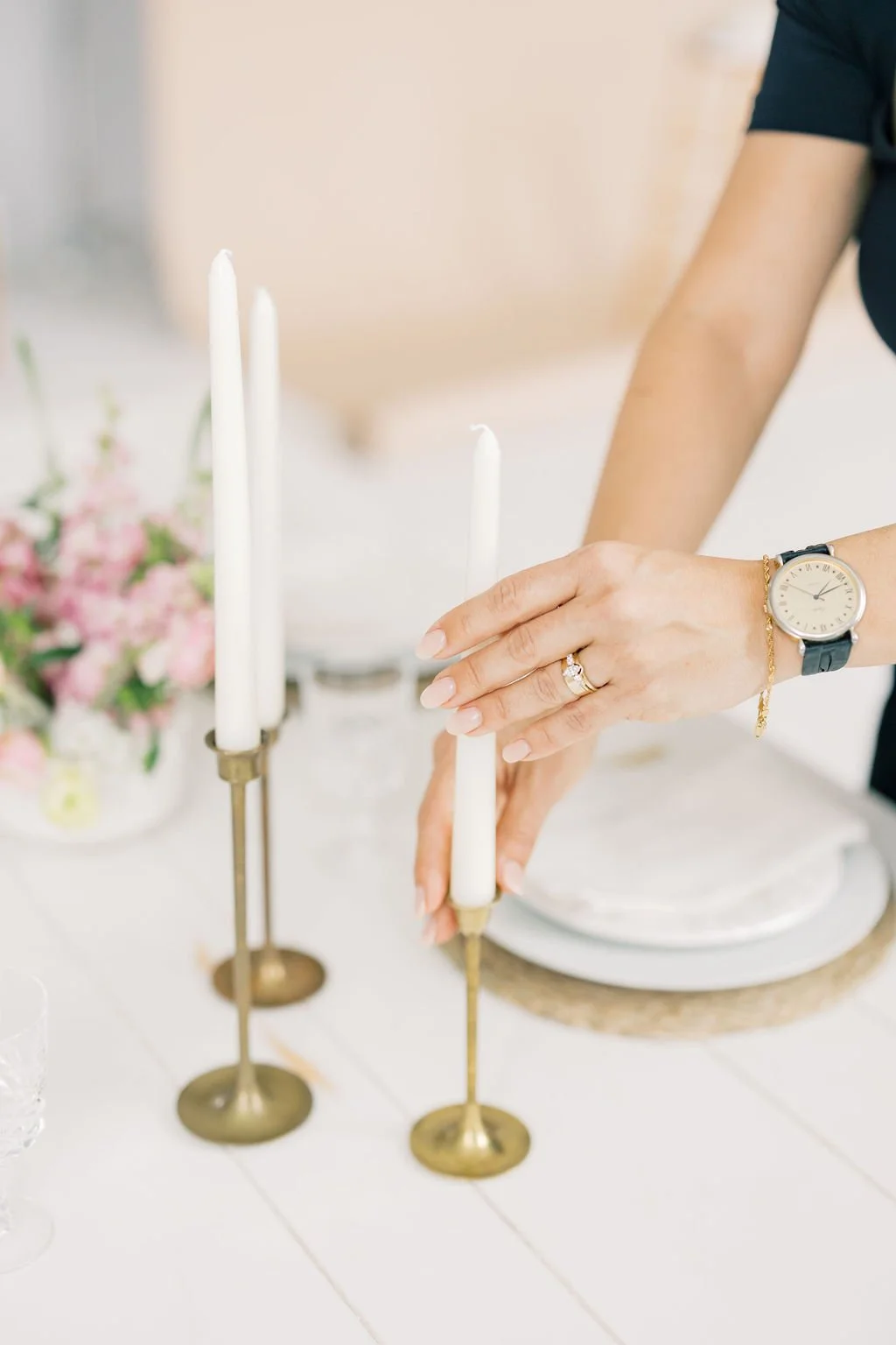 Person decorating a white candle with a gold candle snuffer, wearing a wristwatch and engagement ring, with pink flowers in the background.