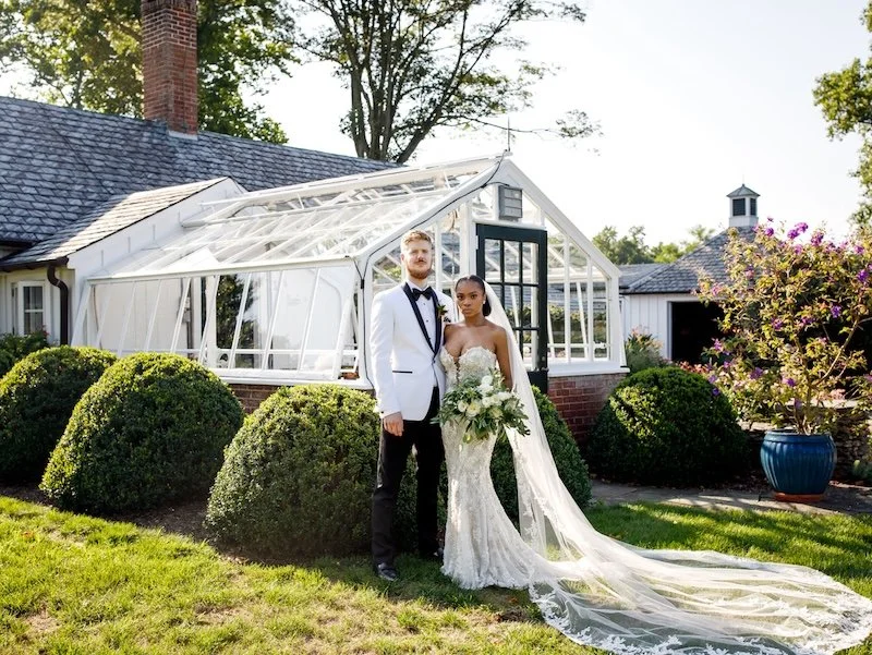 Bride and groom standing outdoors on the lawn of a house with a greenhouse in the background, bride holding a bouquet of white flowers, the groom wearing a white tuxedo jacket.