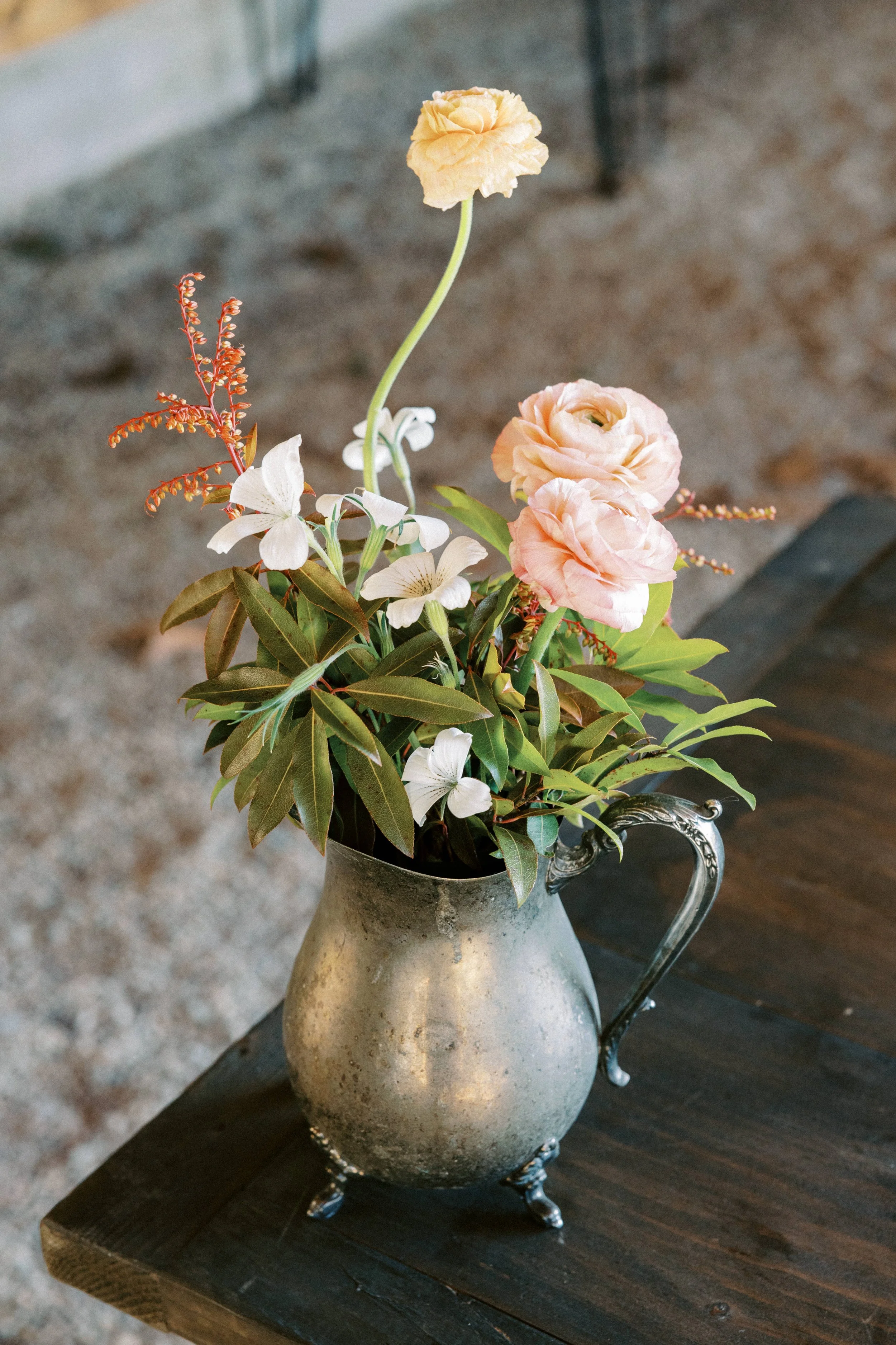A metal silver vase with a handle and three small feet containing a bouquet of pink and cream flowers, including roses and smaller white flowers, on a dark wooden table.