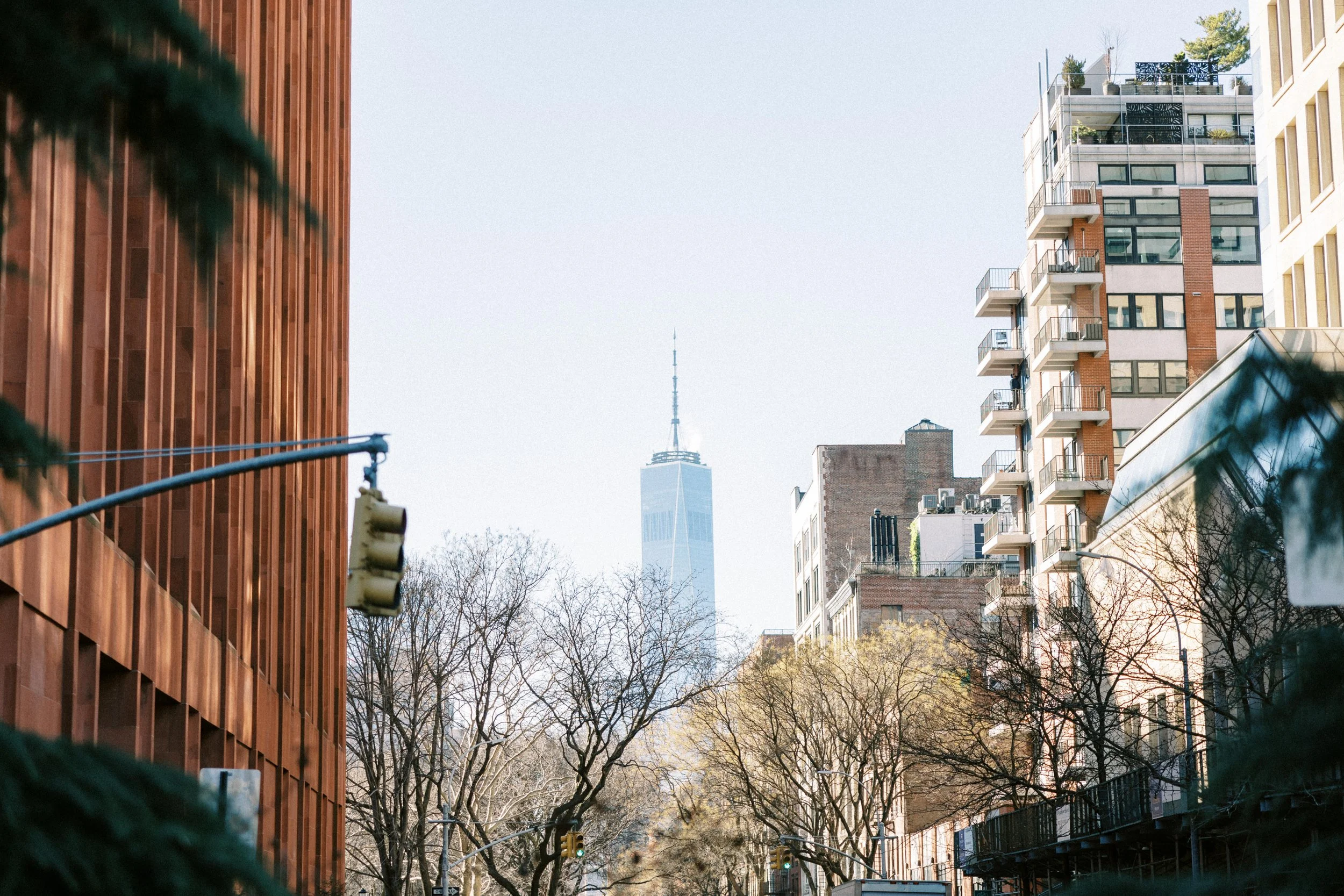 City street view with buildings, trees, traffic lights, and the One World Trade Center in the background.