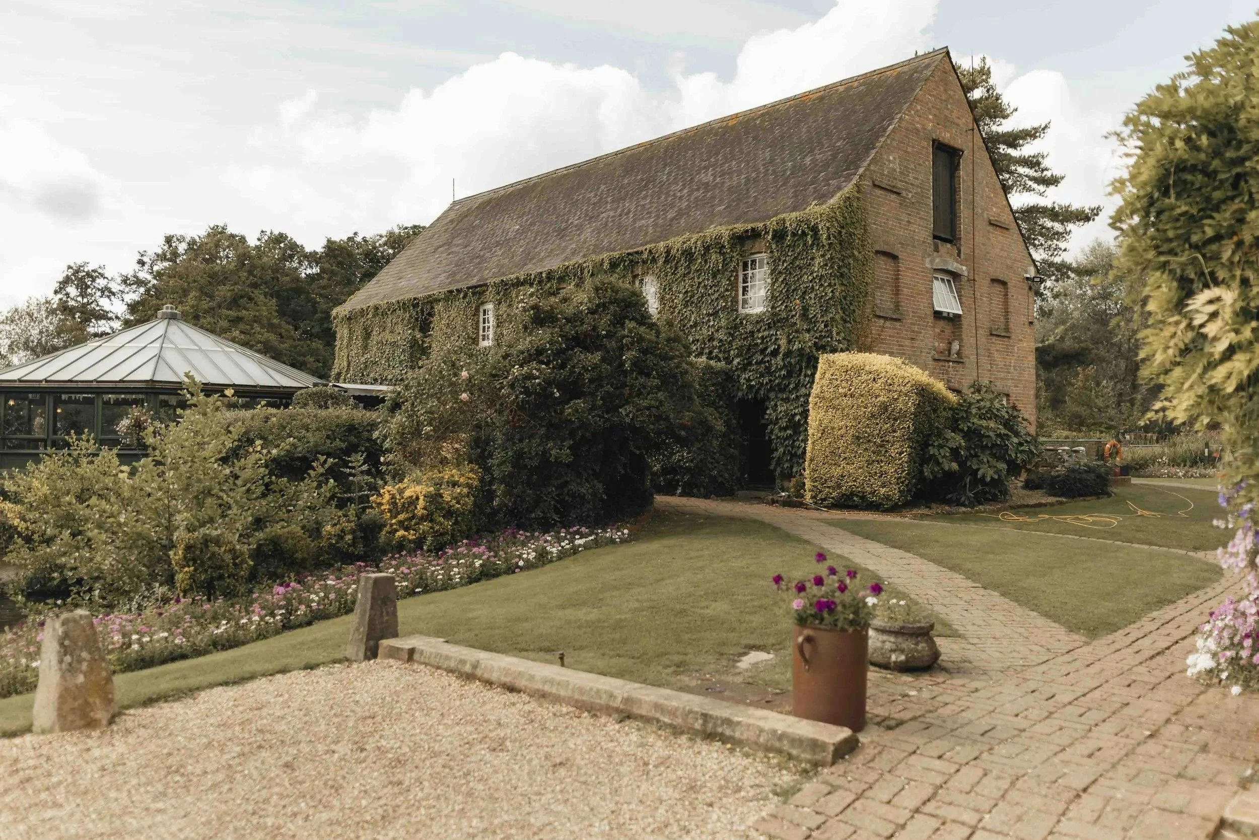 A rustic brick house with ivy-covered walls, surrounded by a well-maintained garden with colorful flowers, a brick pathway, and an outdoor patio area with a glass-roofed garden structure.