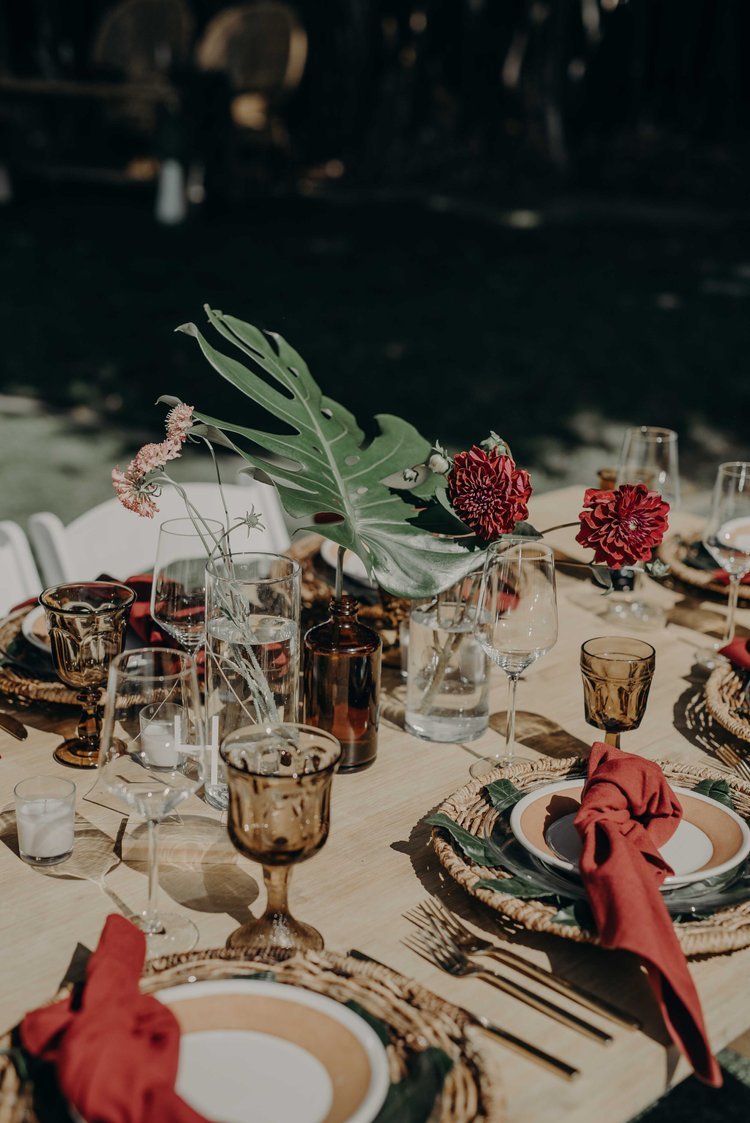 Boho-inspired DIY wedding tablescape with tropical leaves, red napkins, and amber glassware on outdoor reception table