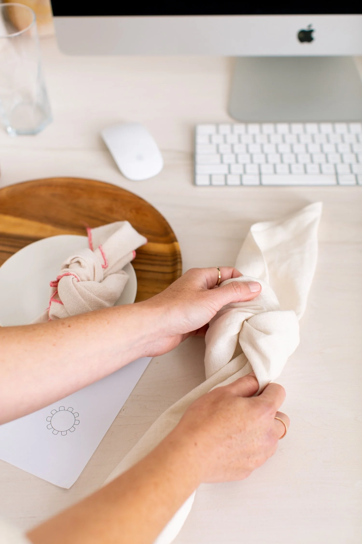 Hands tying a cloth napkin in a knot on a desk with a computer monitor, keyboard, mouse, glass of water, and a round wooden tray.