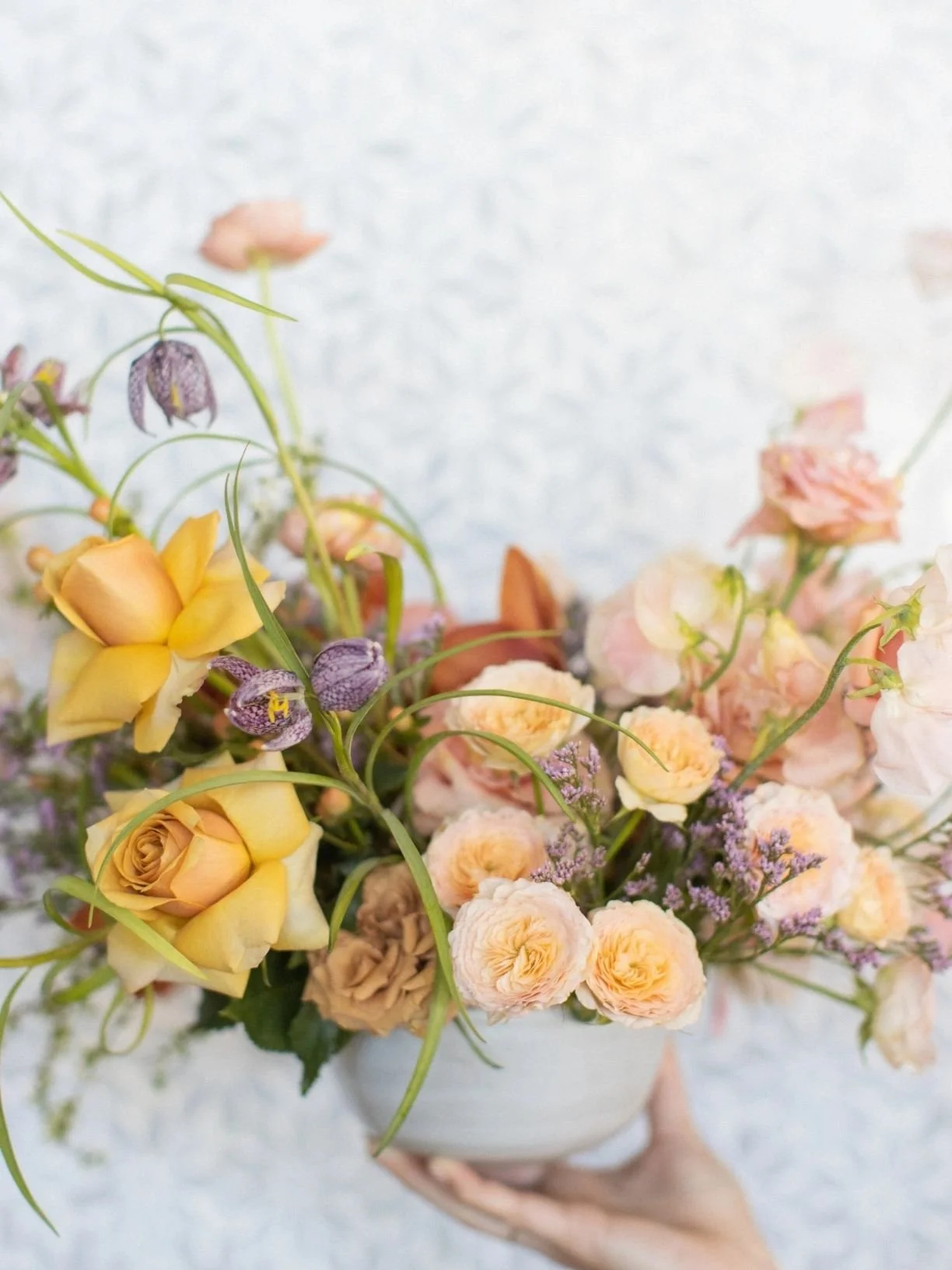 A hand holding a white vase filled with a colorful bouquet of yellow, pink, purple, and peach flowers against a light background.