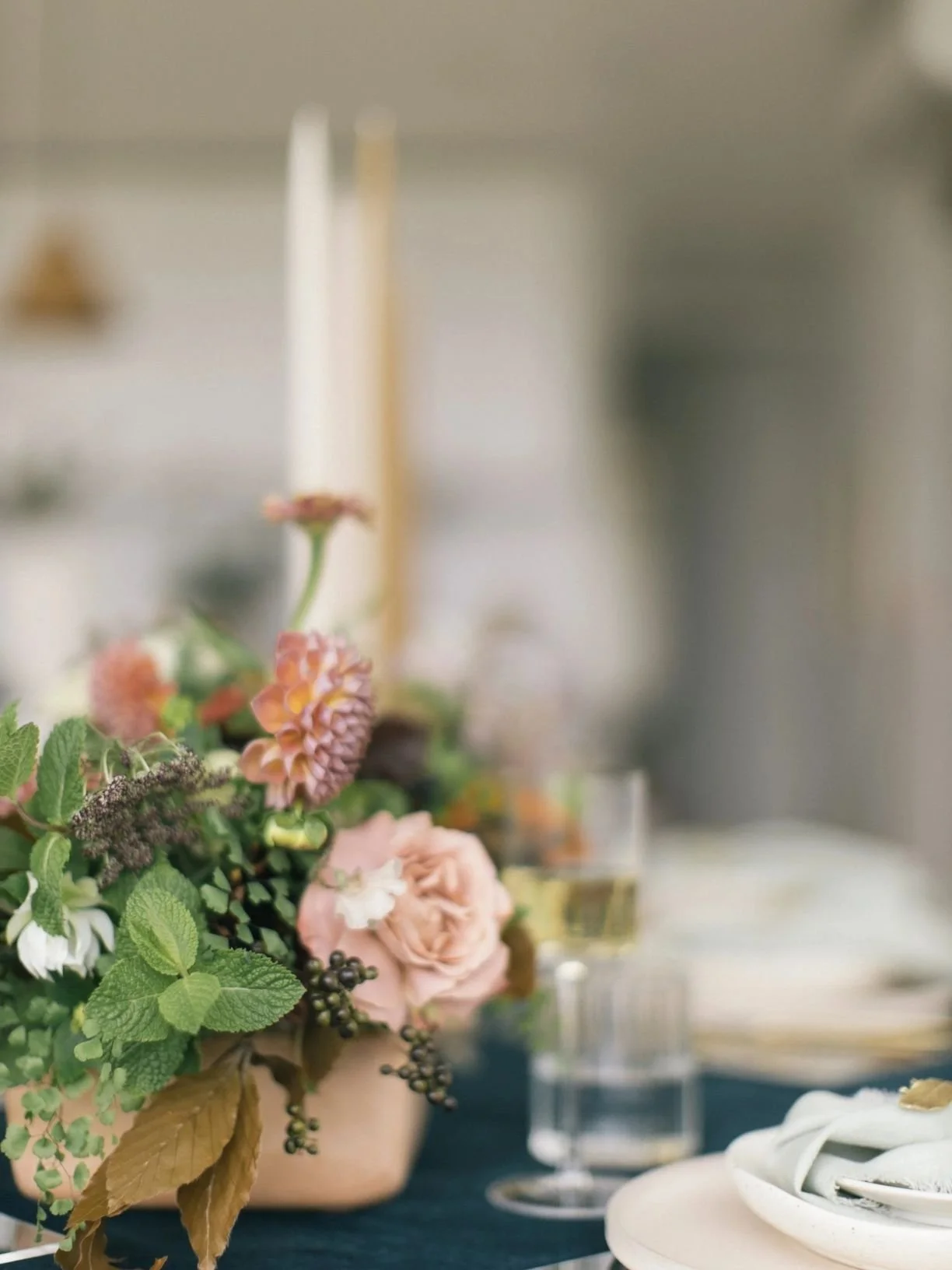 Close-up of a flower arrangement with pink roses, lavender, and greenery on a dining table with a glass of white wine and a white plate in the background.