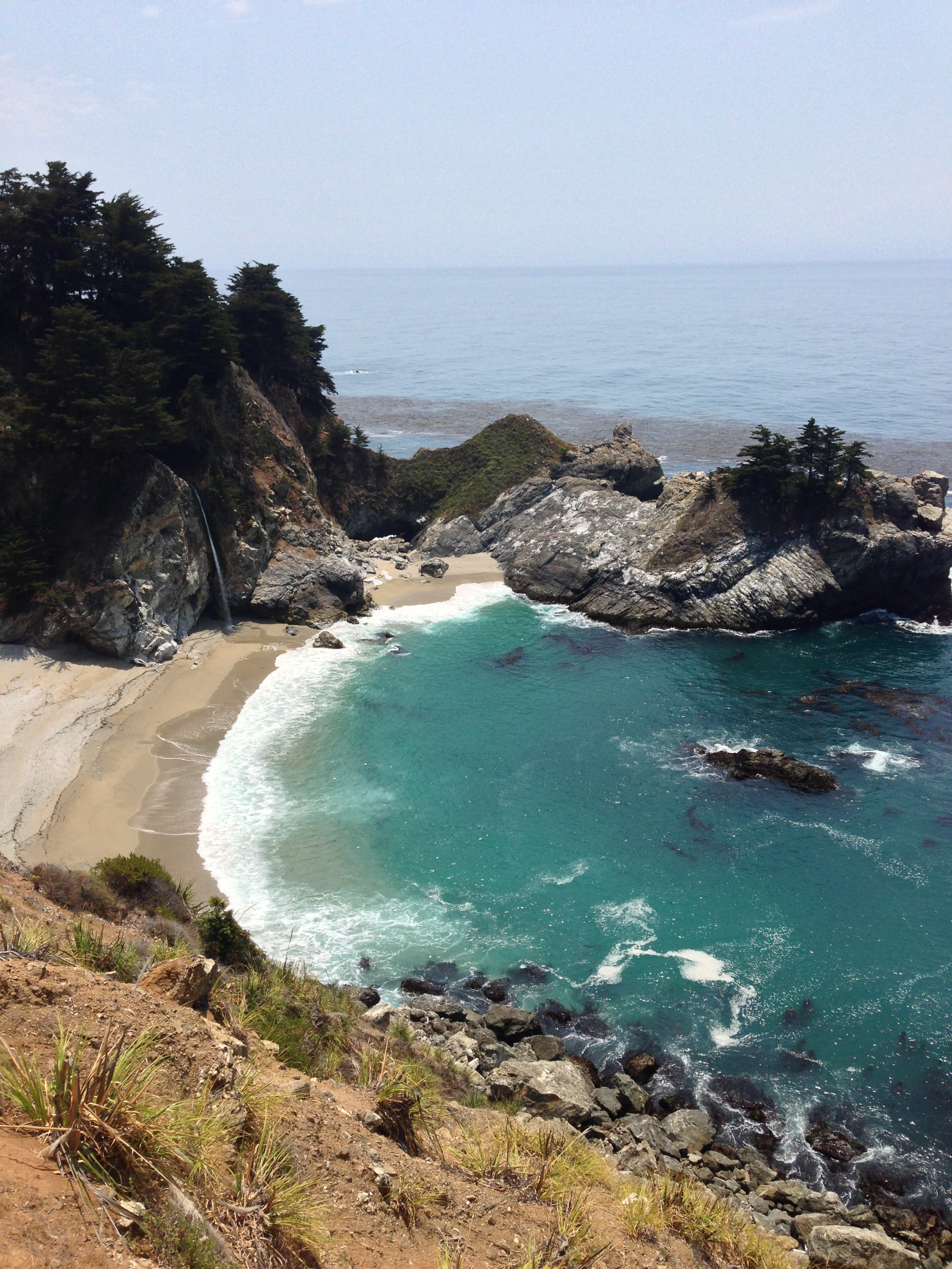 Coastal cove with sandy beach, turquoise water, rocky cliffs, and green trees under a clear sky.