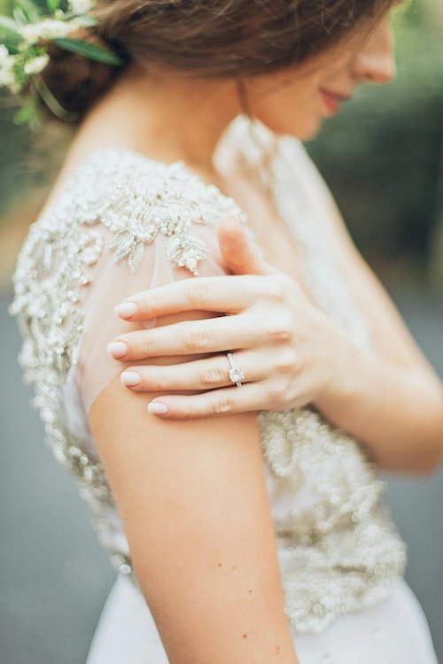 Close-up of a woman in a wedding dress showing her hand with an engagement ring on her ring finger, gently touching her shoulder.