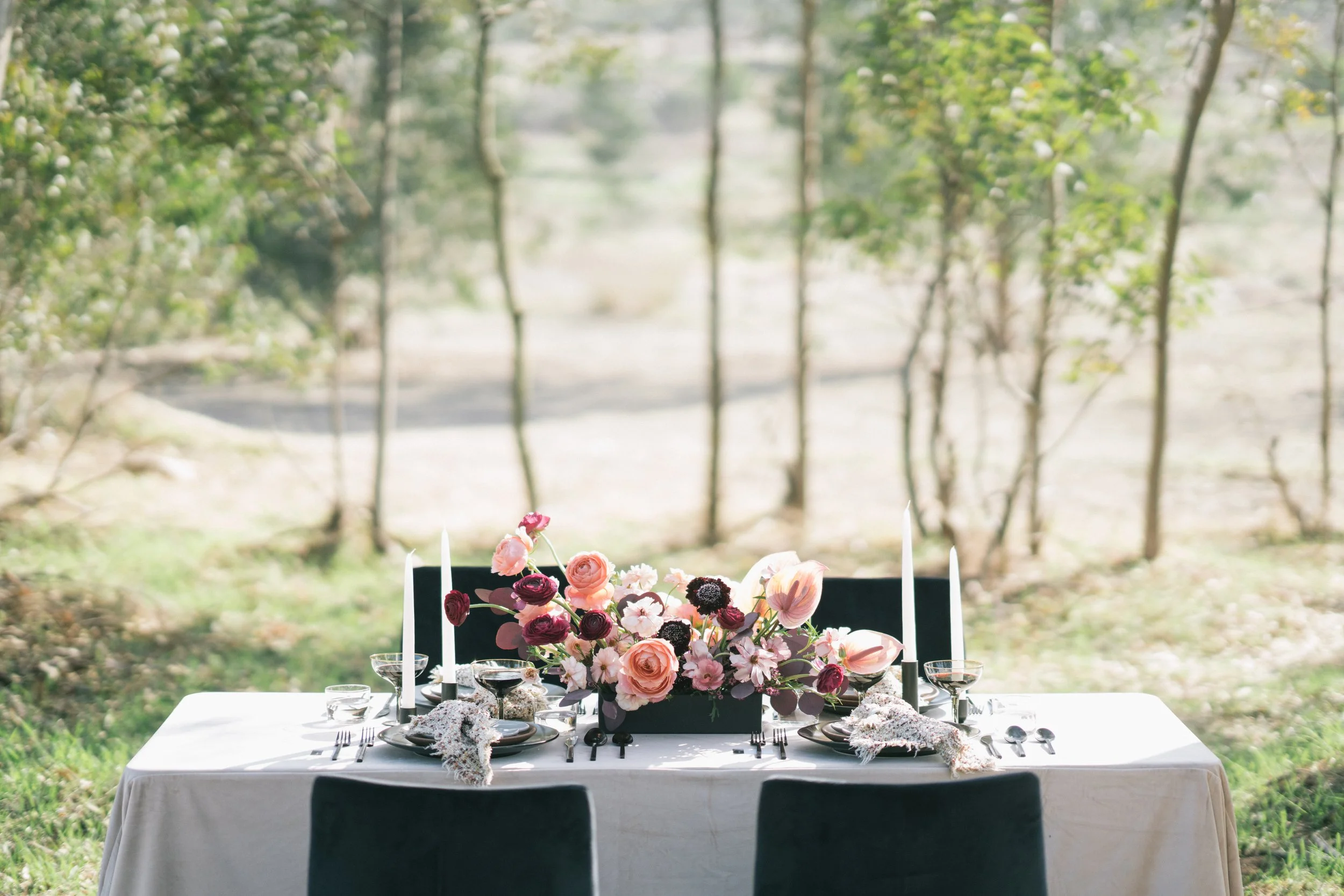 Outdoor wedding table styled with pink and burgundy flowers, black chairs, taper candles, and white linens.