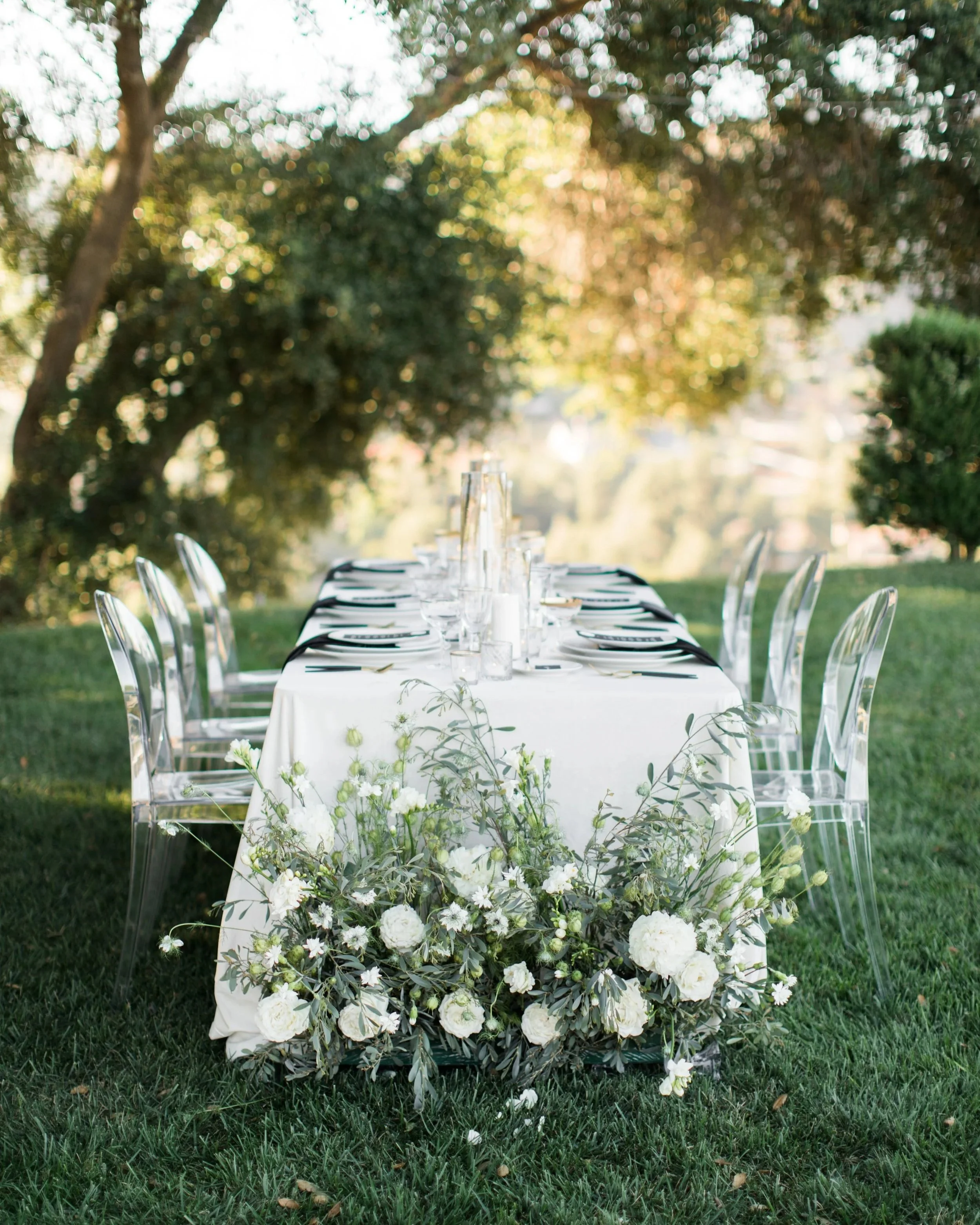 Elegant outdoor wedding table with black napkins, clear ghost chairs, and white florals under tall trees in the Hudson Valley