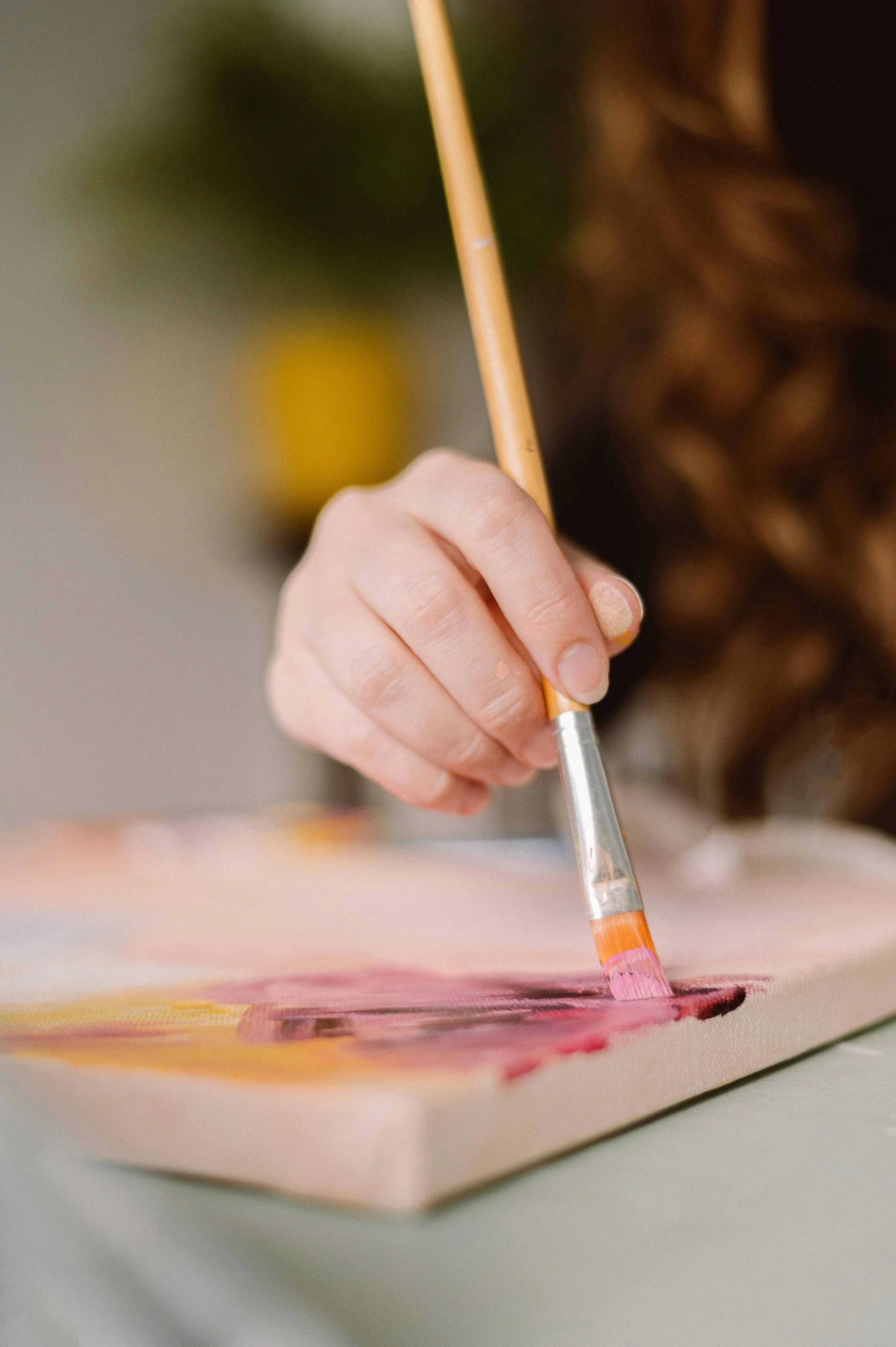 Close-up of a person's hand holding a paintbrush and painting on a canvas with pink and yellow colors.