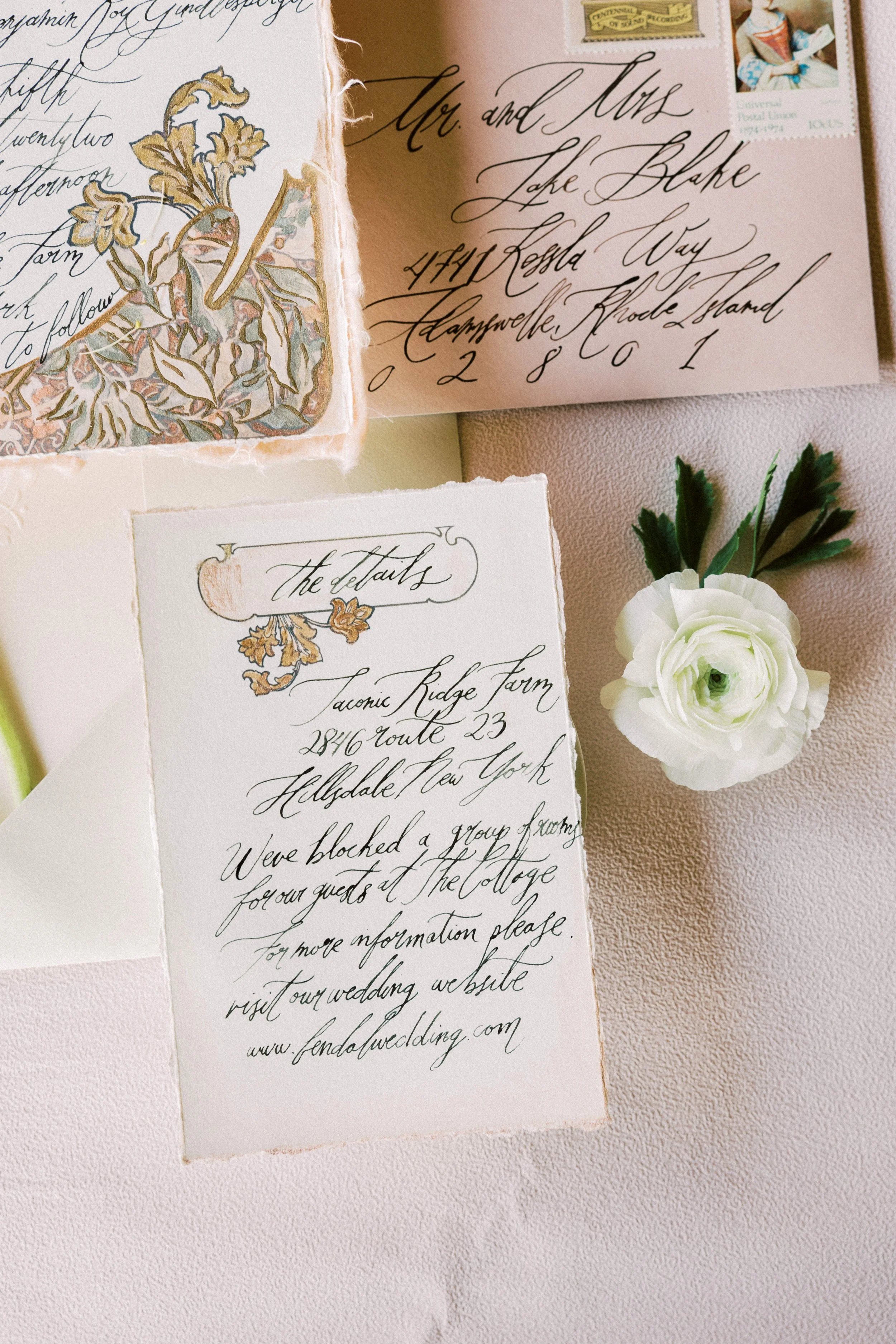 A handwritten wedding invitation with elegant calligraphy, decorated with floral illustrations and a white ranunculus flower with greenery, placed on a textured pink surface.