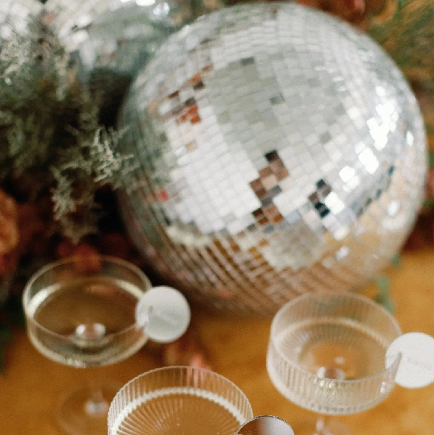 A disco ball with small reflective tiles is hanging, reflecting light. Surrounding it are empty small glass candle holders with white candles inside, placed on a wooden surface. There are some greens on the left side of the image.