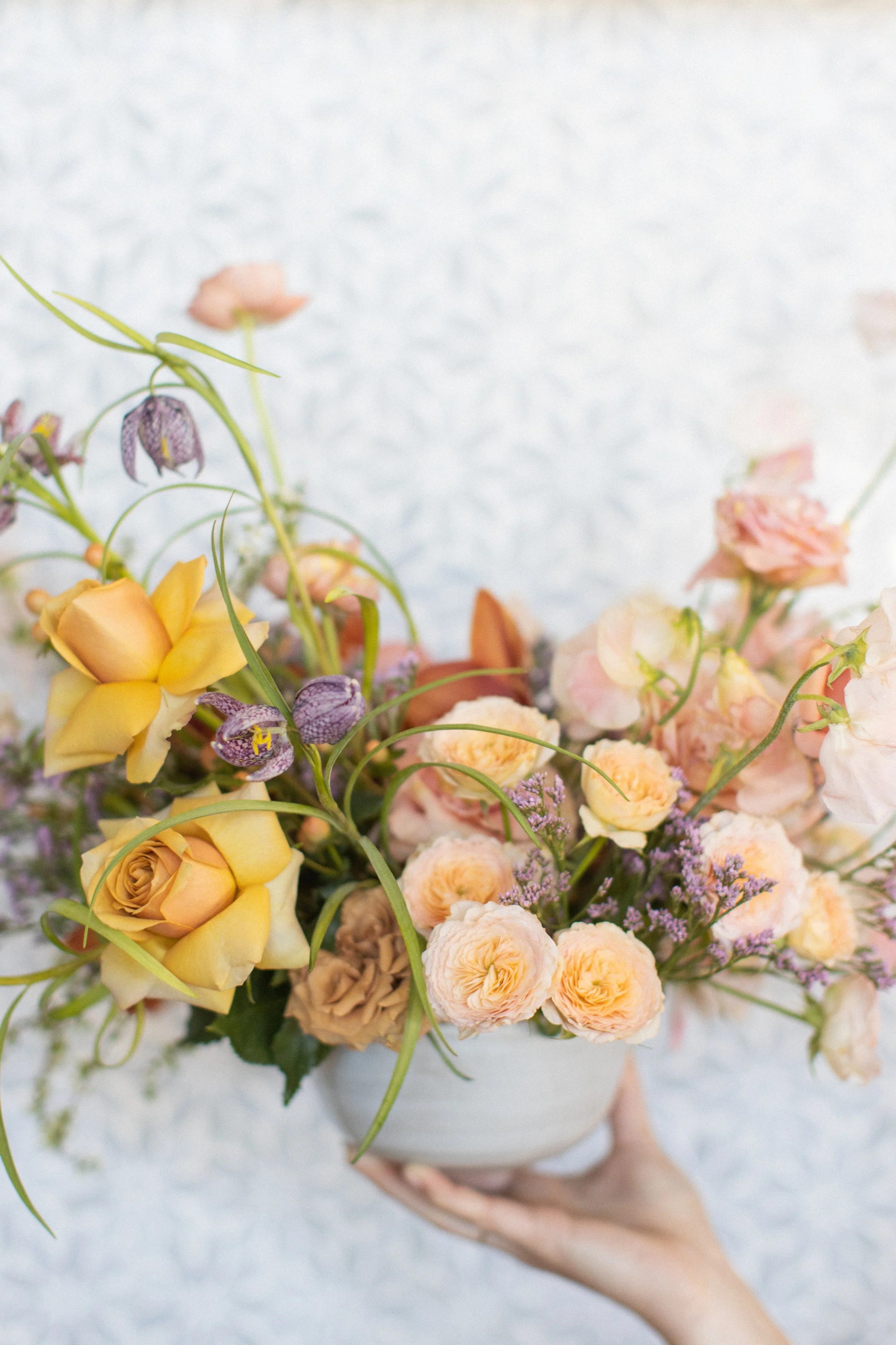 A hand holding a bowl of assorted flowers including yellow roses, pink blooms, purple flowers, and greenery against a light background.