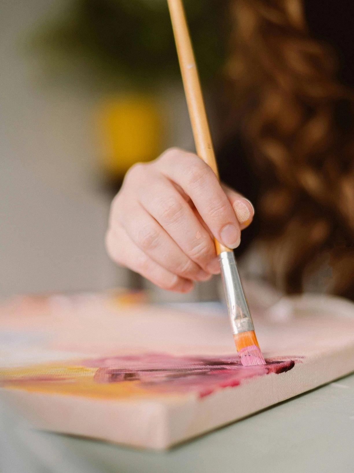 Close-up of a person holding a paintbrush, painting with pink and dark colors on a canvas.
