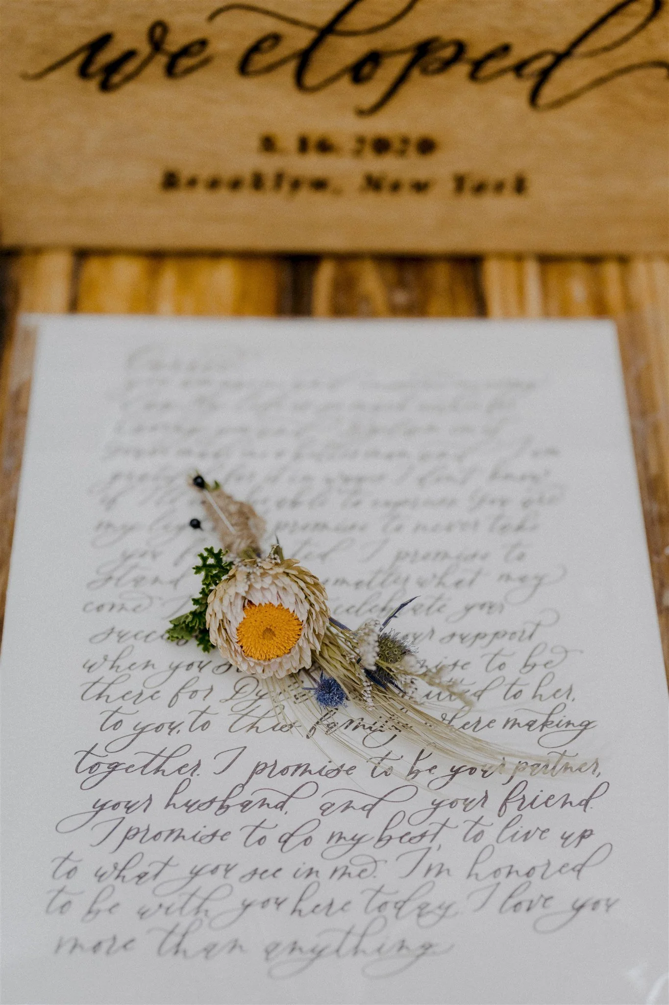 A vintage-style wedding vow letter with cursive handwriting, decorated with a small floral boutonniere consisting of a cream-colored flower, yellow center, green leaves, and dried blue and purple flowers.