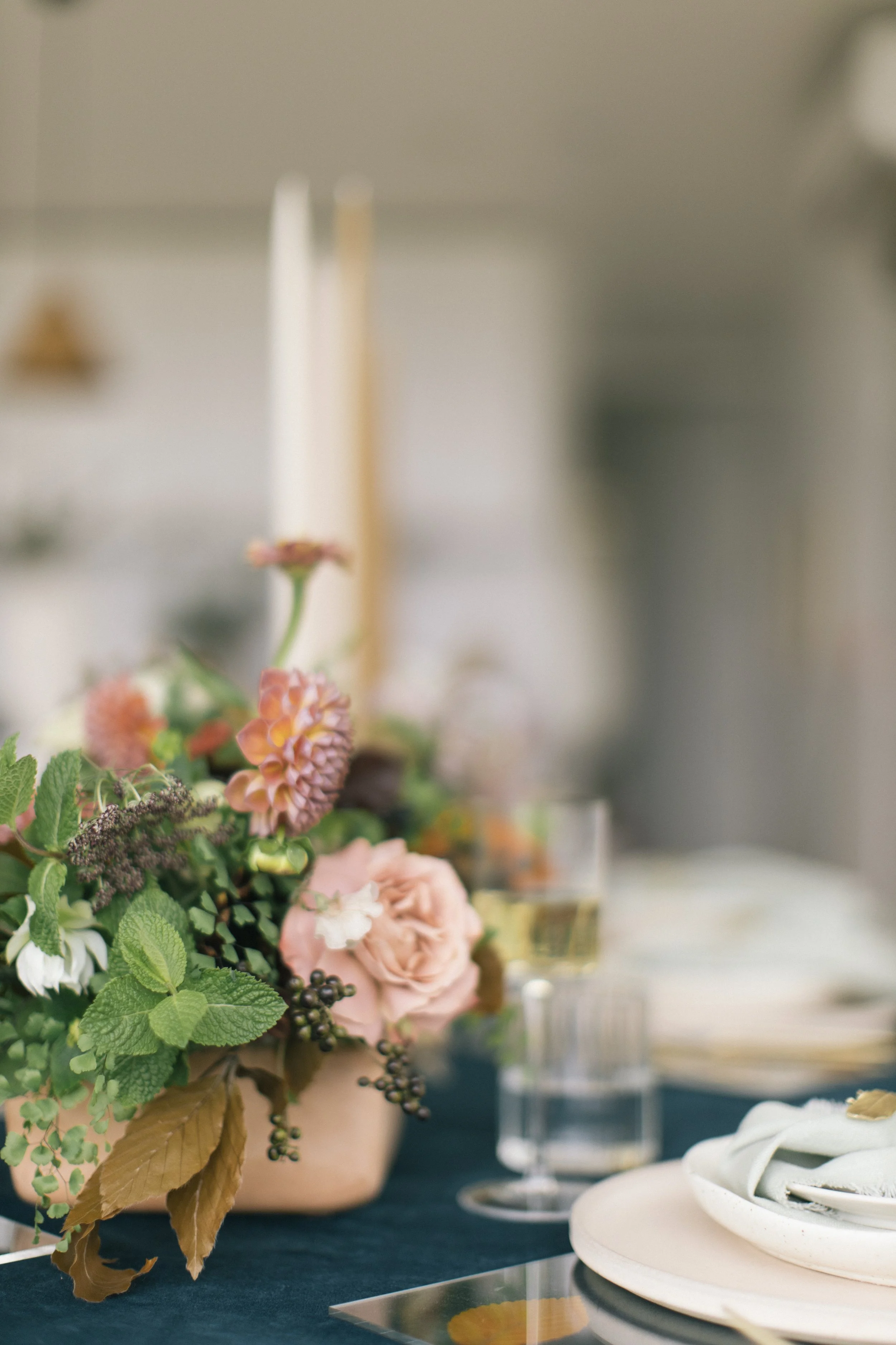 Close-up of a floral centerpiece with pink roses, dahlias, greenery, and berries on a dining table with a glass of white wine and place setting.
